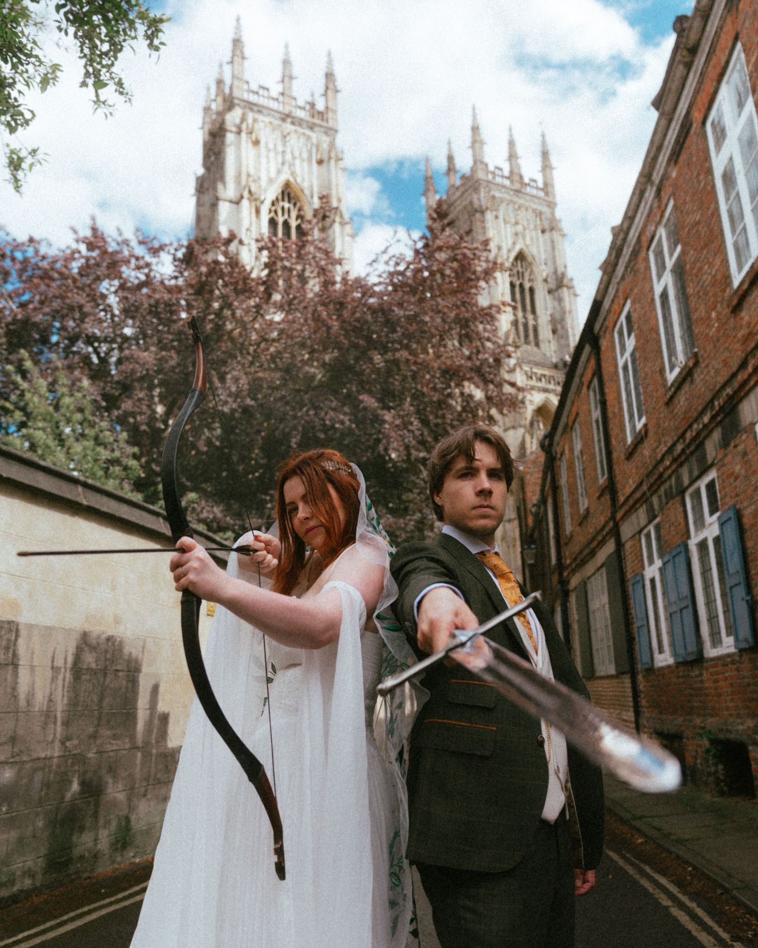 Medieval-themed wedding couple pose back-to-back with bow and arrow near Gothic cathedral in York.