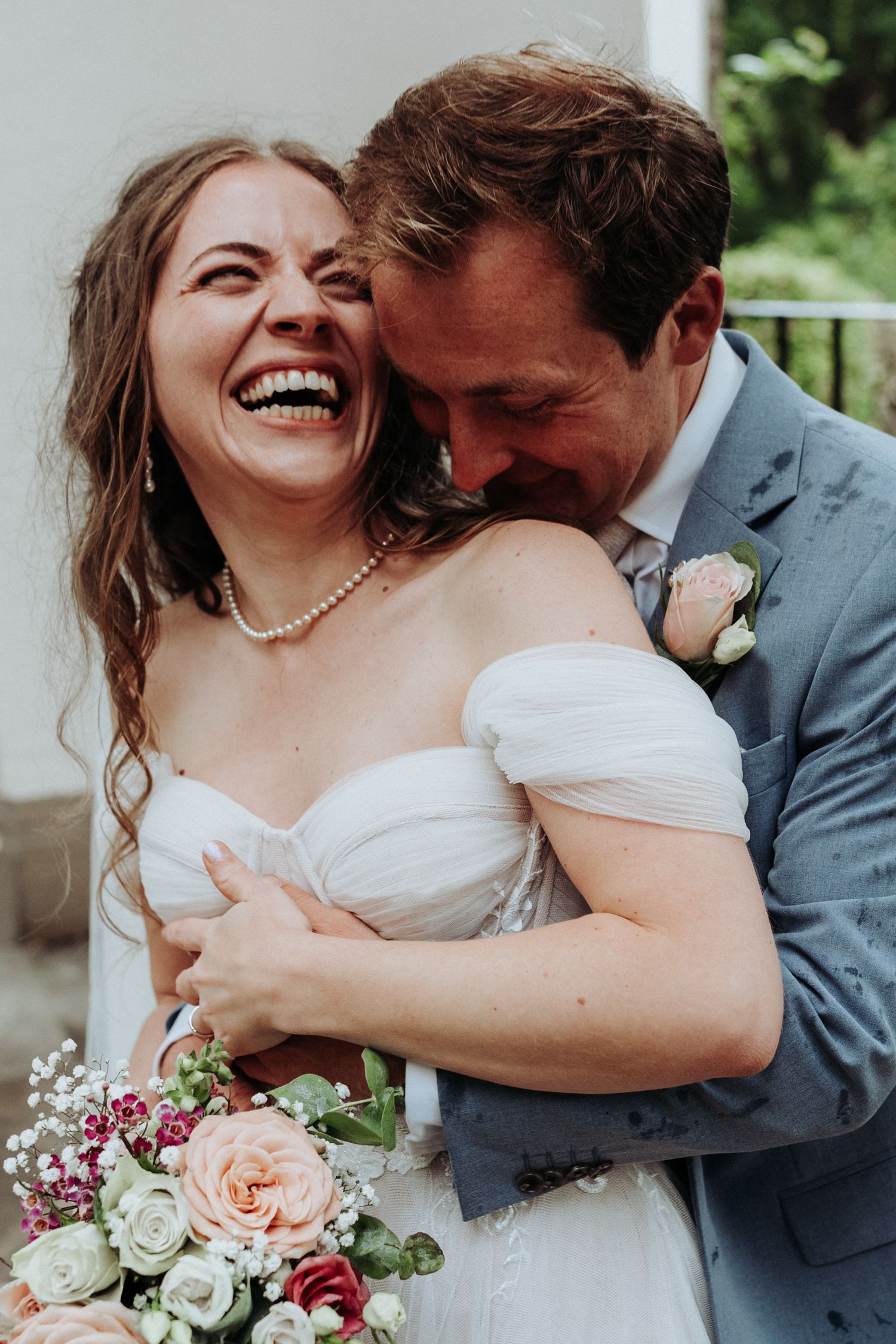 Groom hugs bride from behind as she laughs joyfully, holding a pastel rose wedding bouquet.