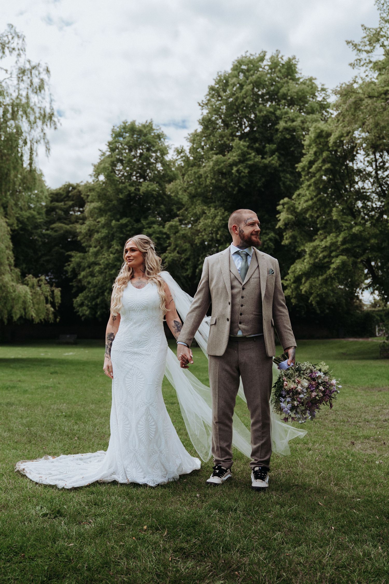 Bride and groom holding hands walking through lush green park, bride in lace gown with veil, groom in tan suit.