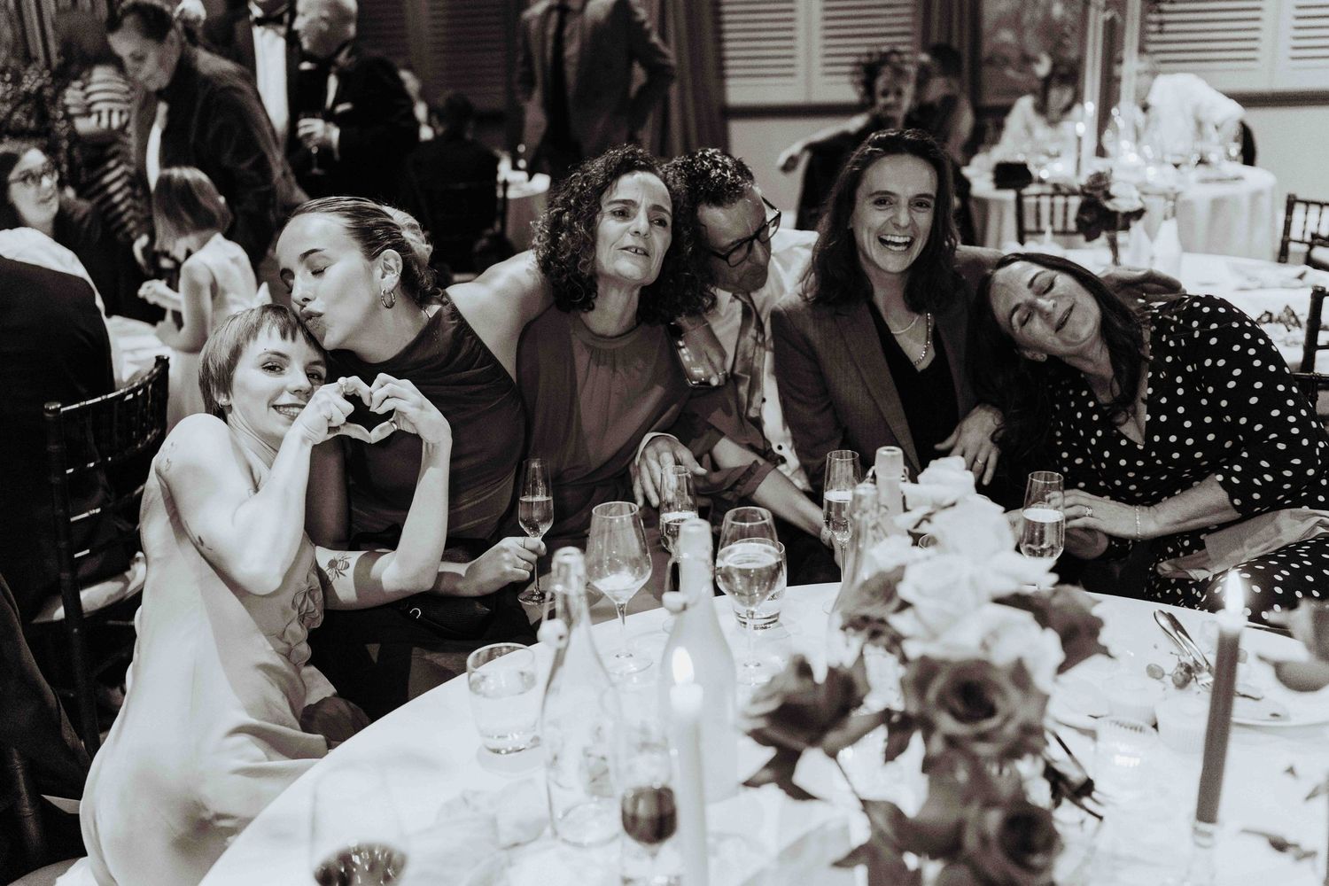 Group of wedding guests laughing and posing together at a reception dinner table in black and white.