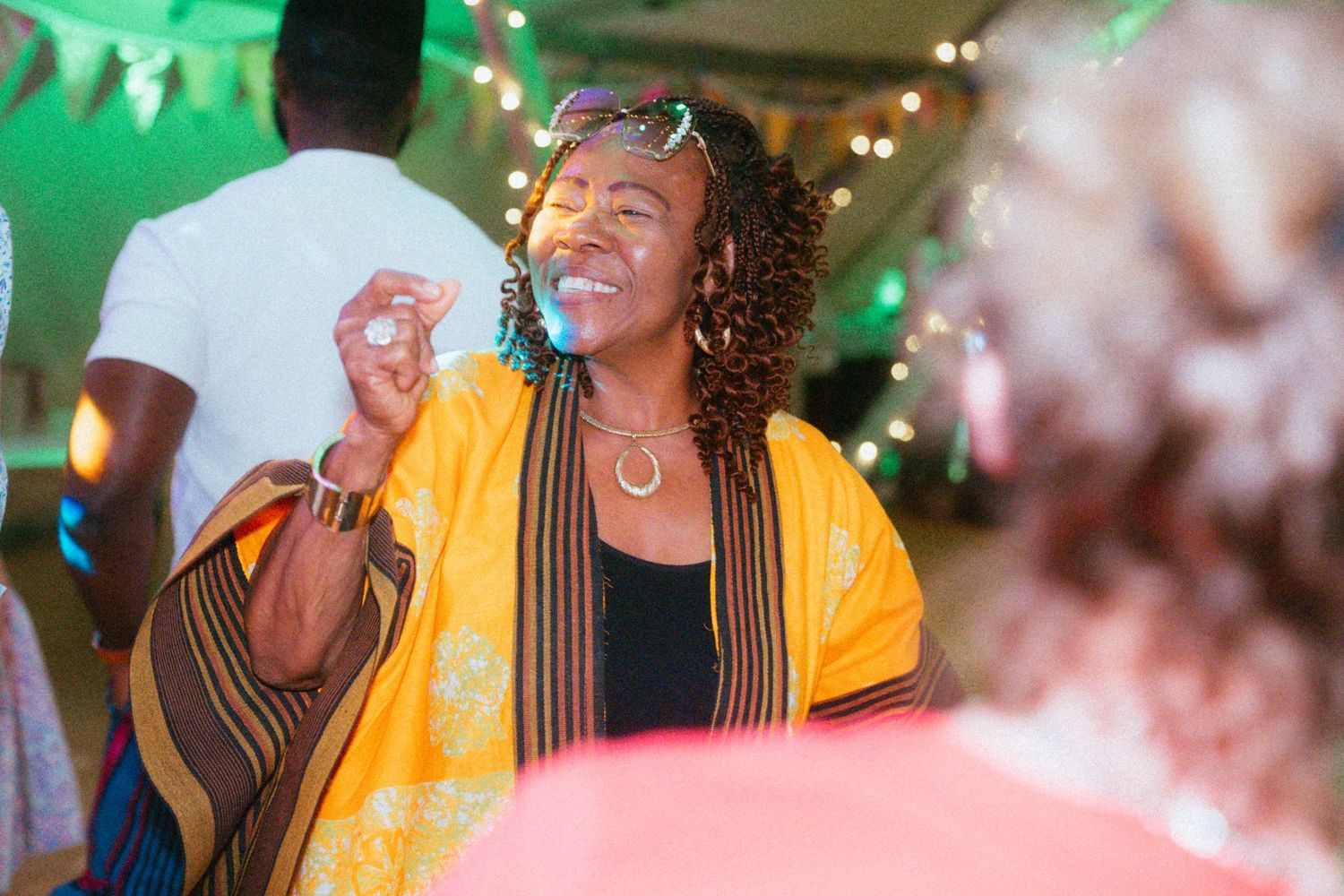 Woman in vibrant yellow African print wrap dancing joyfully at an outdoor evening celebration with string lights.
