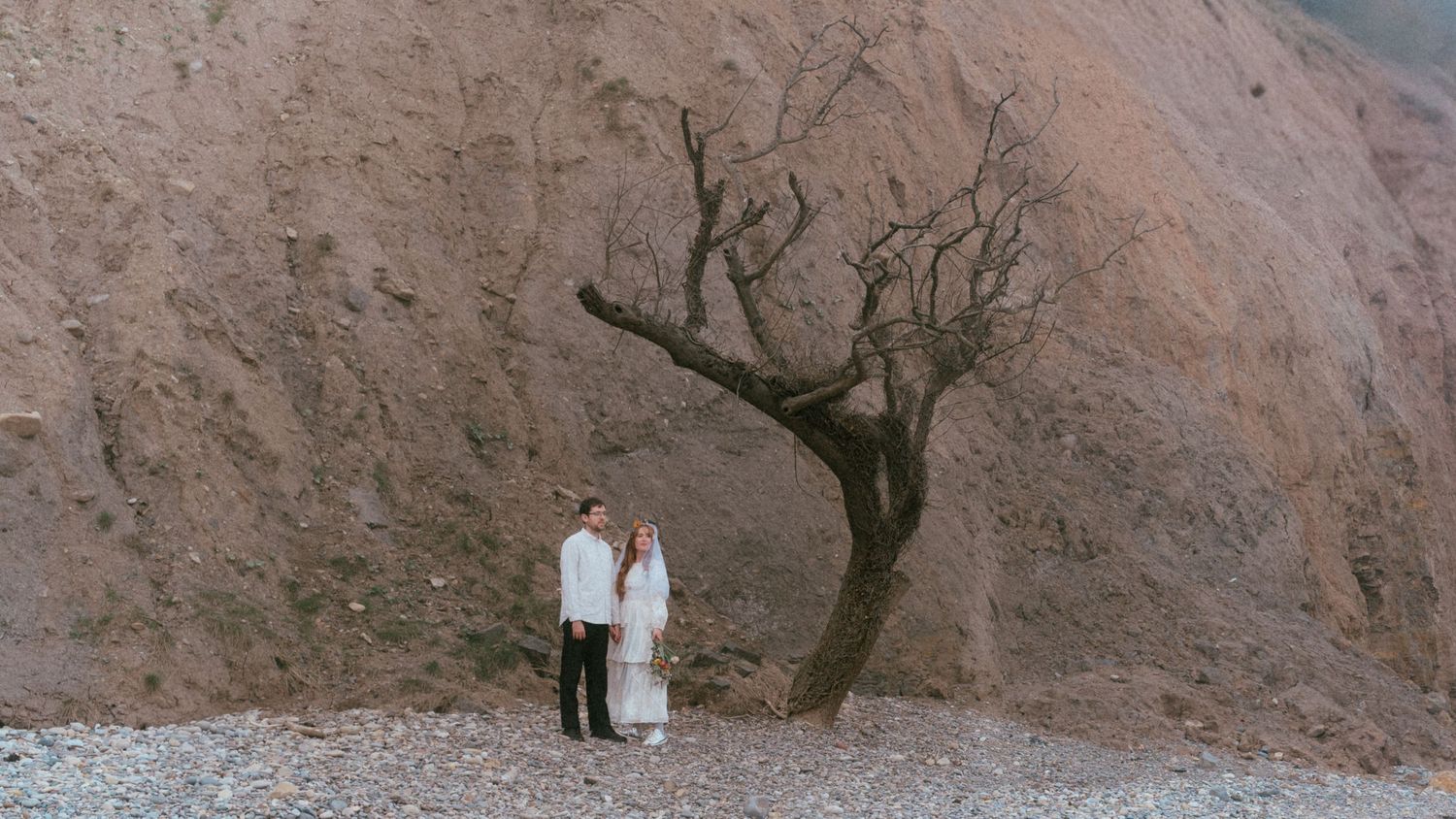 Couple in white attire standing beneath a bare tree against a dramatic rocky mountain backdrop.
