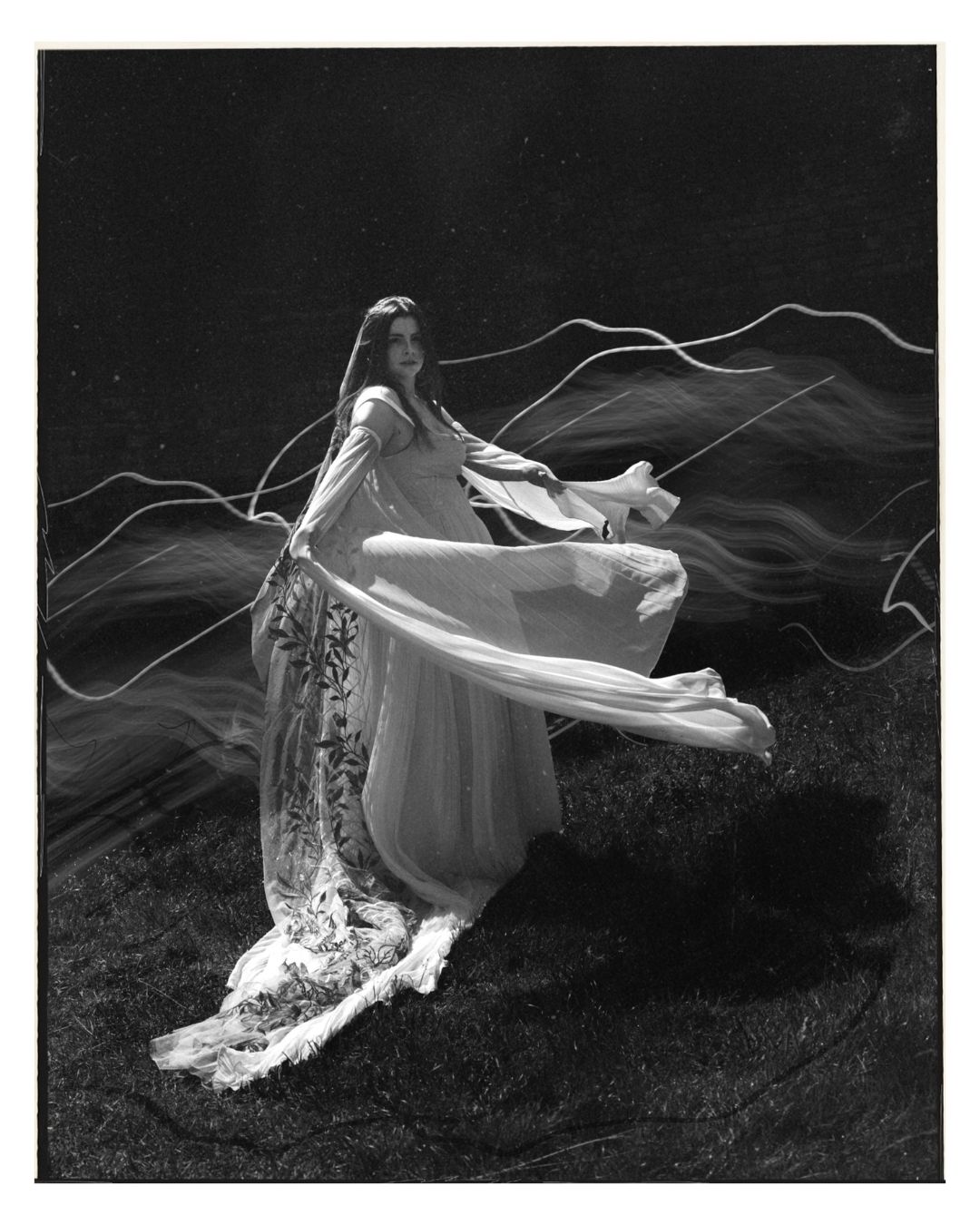 Black and white photo of a woman in a flowing white gown with light trails swirling around her outdoors.
