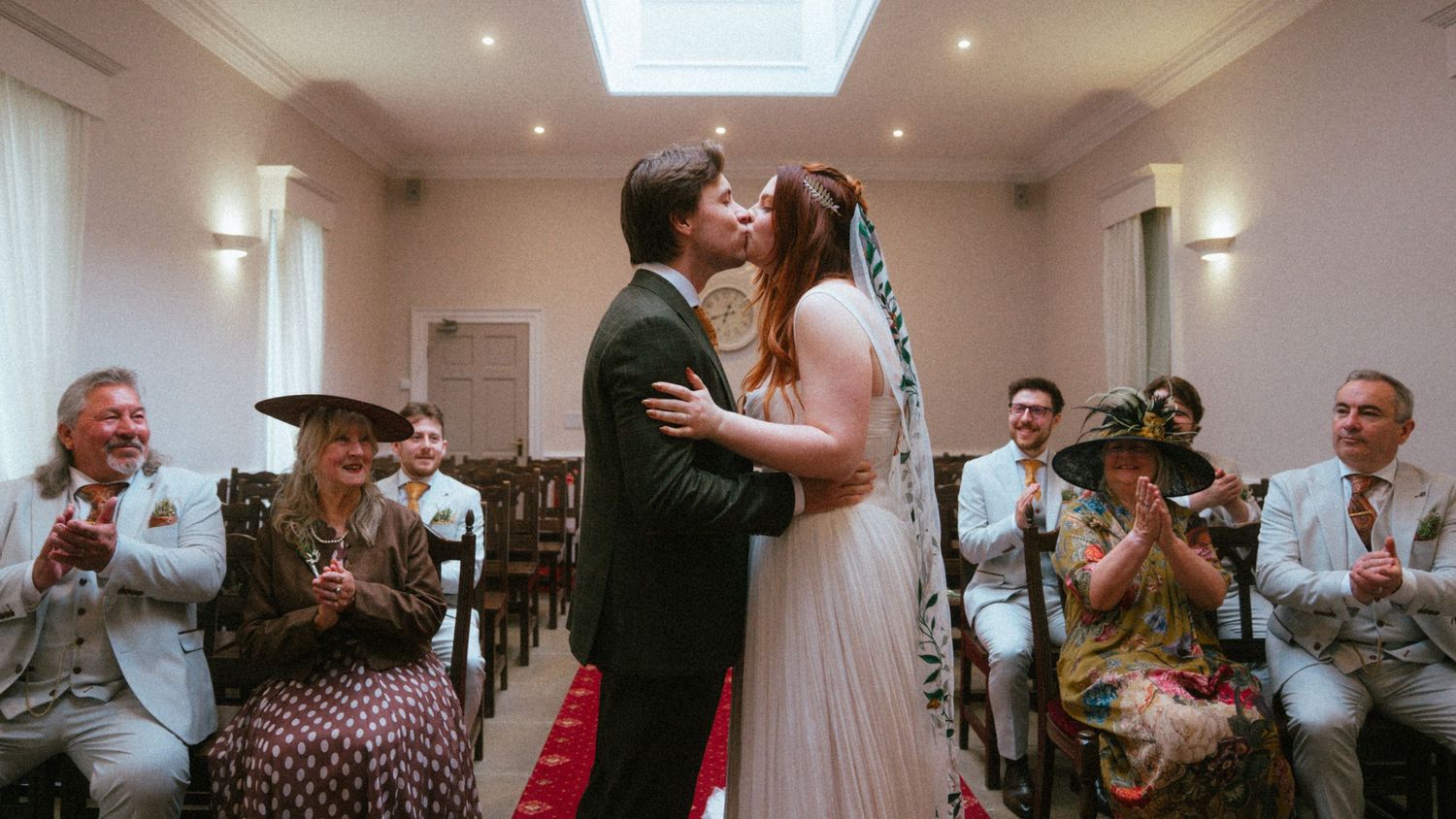 Newlywed couple shares first kiss at wedding ceremony while guests applaud in elegant white venue.