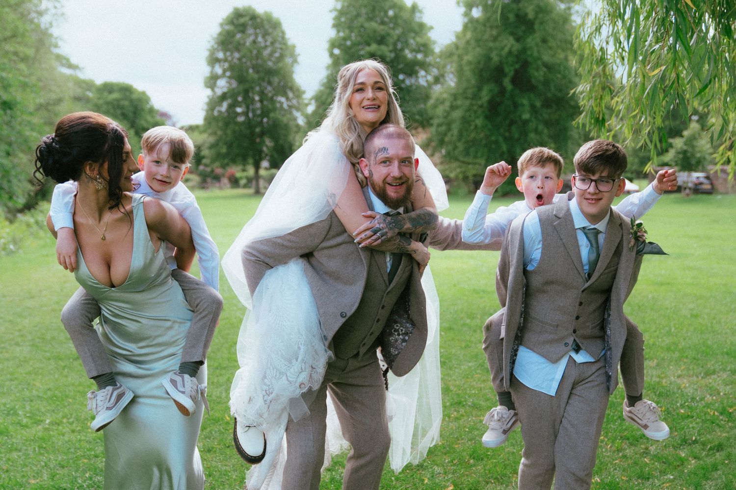 Laughing wedding party with bride piggyback on groom celebrating on scenic green lawn.