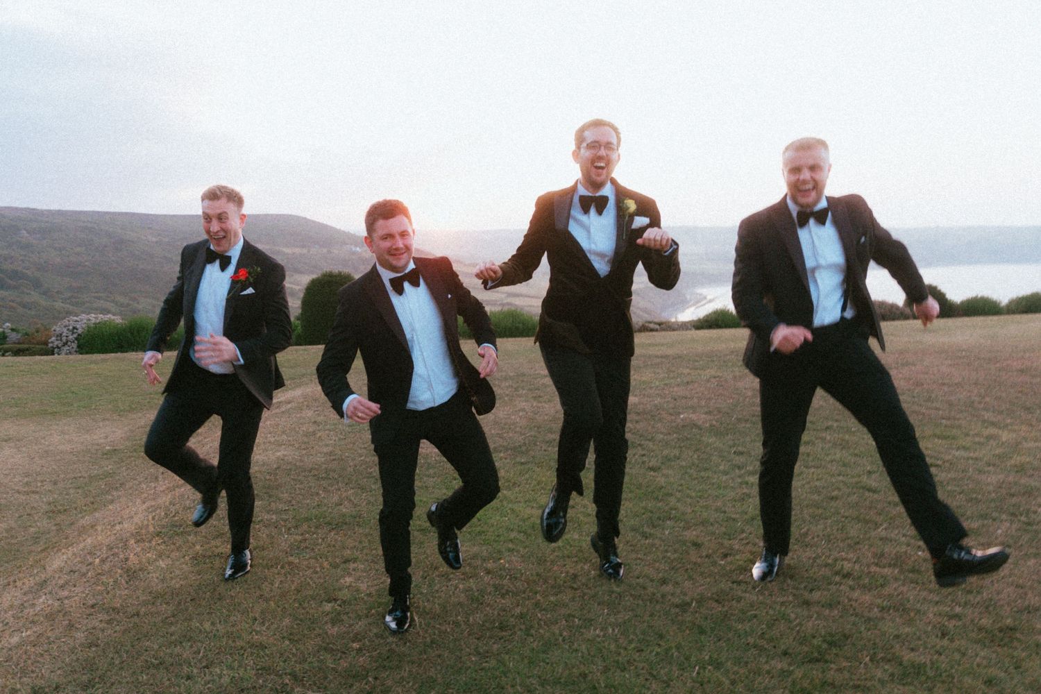 Four groomsmen in black tuxedos and bow ties jump in sync on a grassy hillside overlooking the sea at sunset.