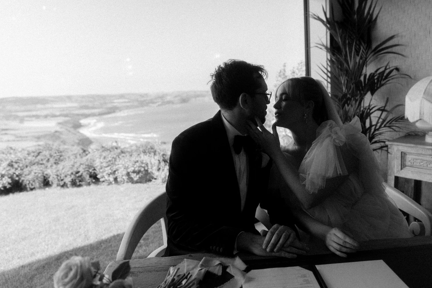 Black and white photo of a couple in formal attire sharing an intimate moment at a table with a scenic ocean coastline behind them.