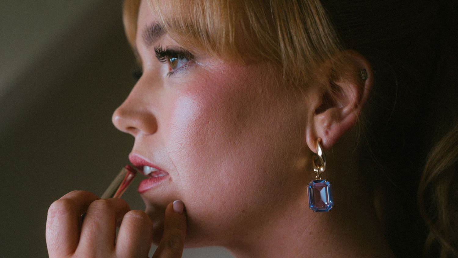 Side profile of a woman with blonde bangs applying lip liner while wearing a sparkling blue gemstone drop earring.