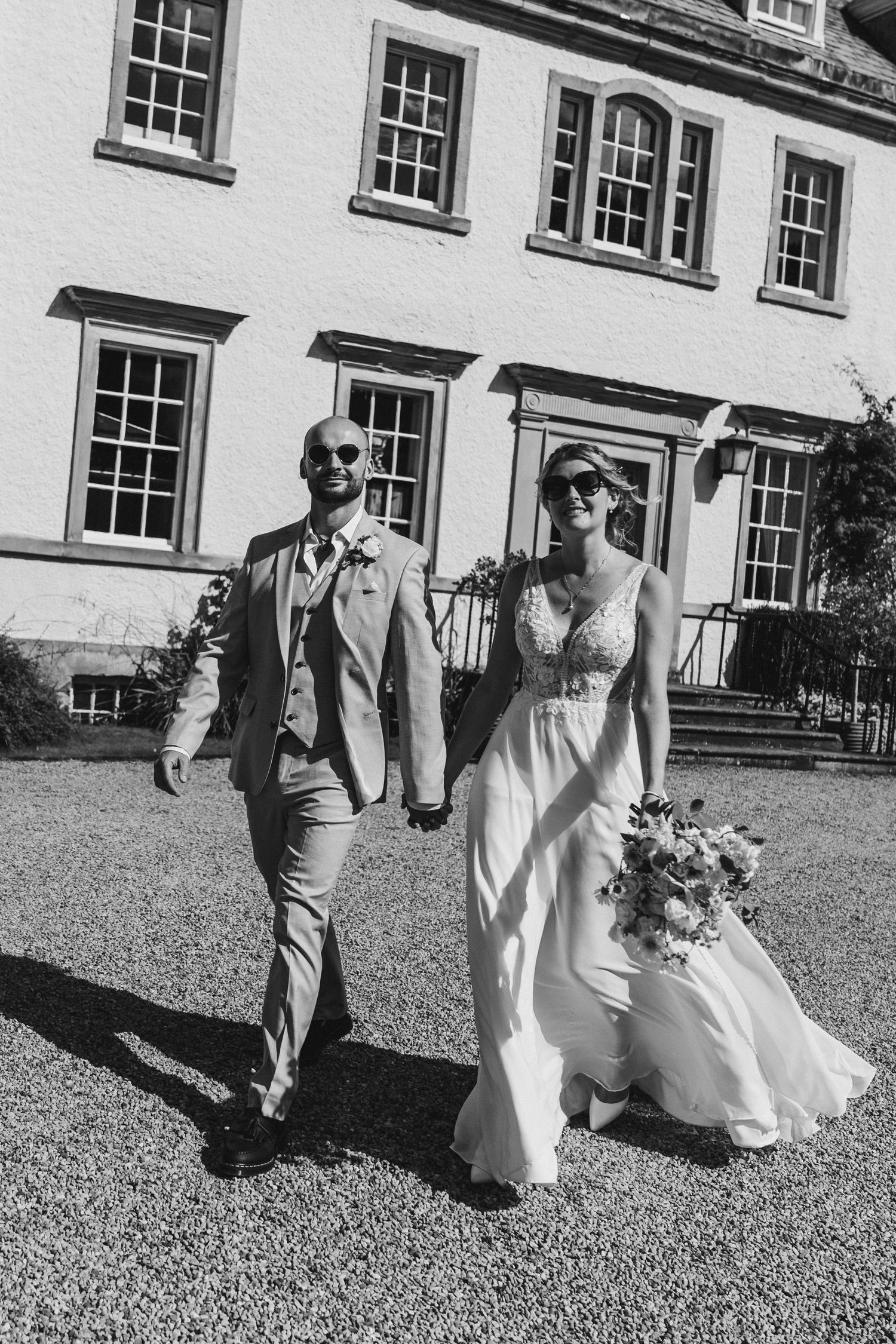Bride and groom walk hand in hand wearing sunglasses outside a grand manor house.