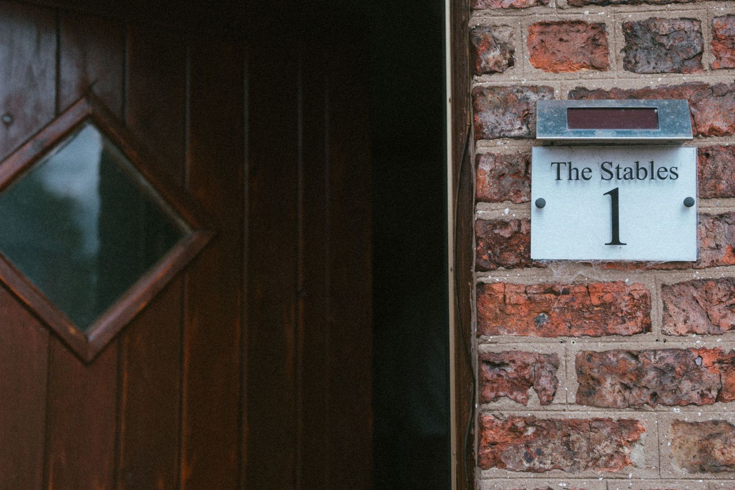 Brick wall with a white house number sign reading The Stables 1 next to a dark wooden door.