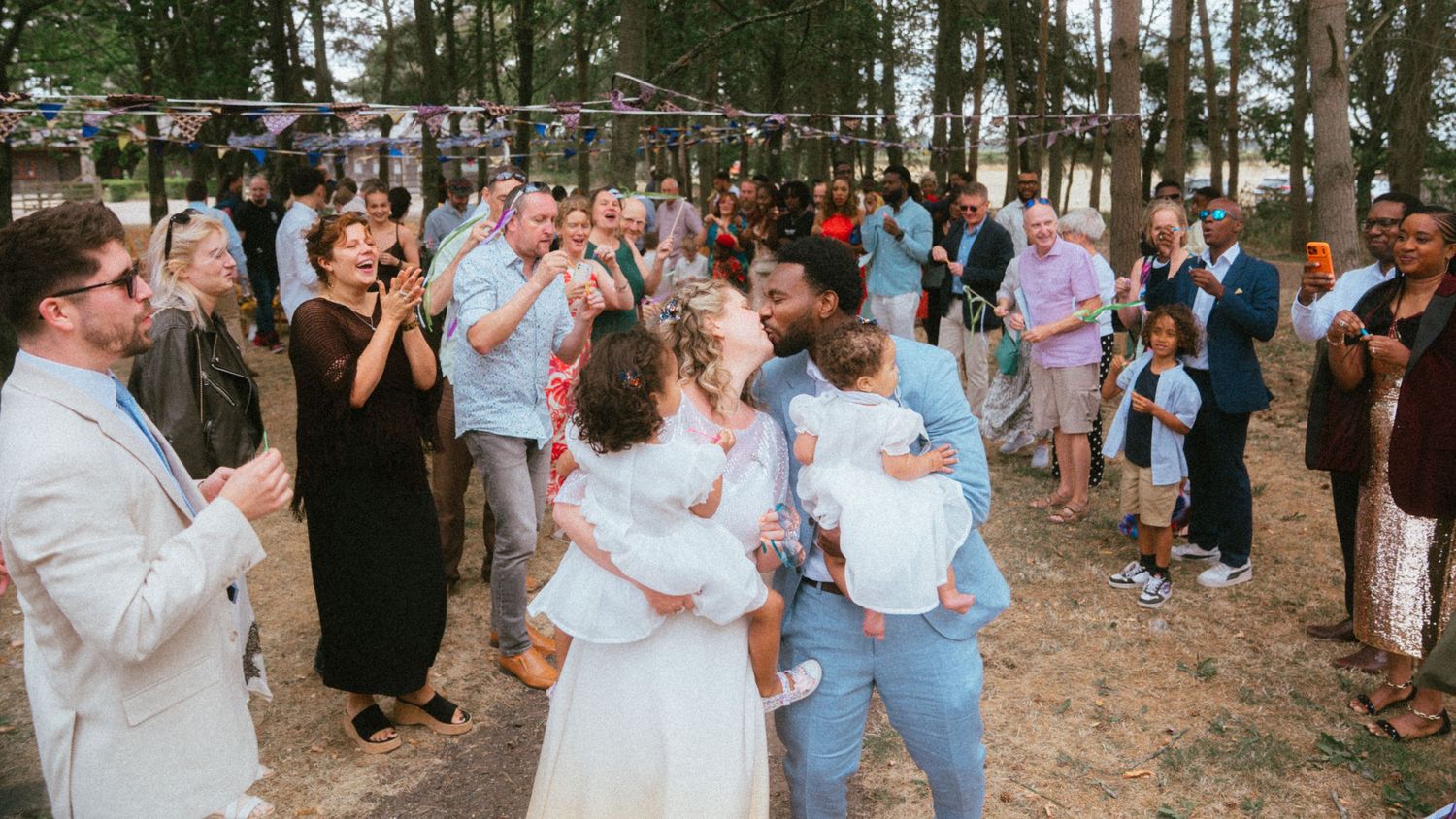 Happy wedding couple embracing with guests celebrating at a joyful outdoor forest wedding reception.