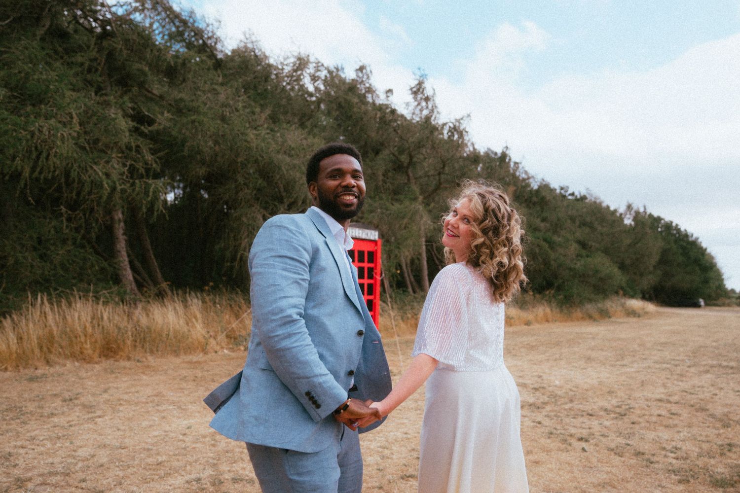 Interracial couple holding hands in a field, groom in light blue suit and bride in white dress with red phone box.
