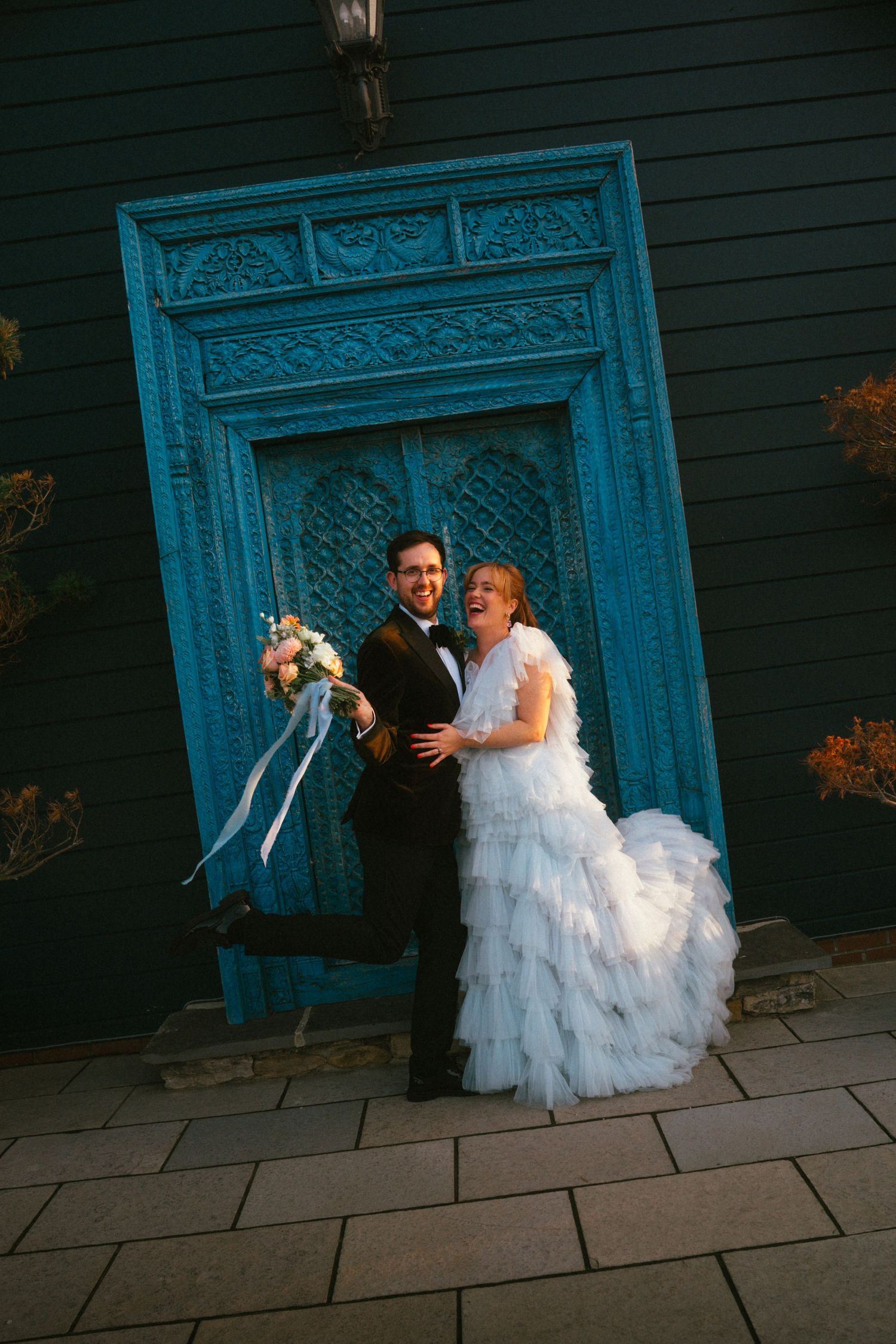 Joyful bride and groom posing in front of ornate teal door, bride in ruffled white gown holding bouquet.