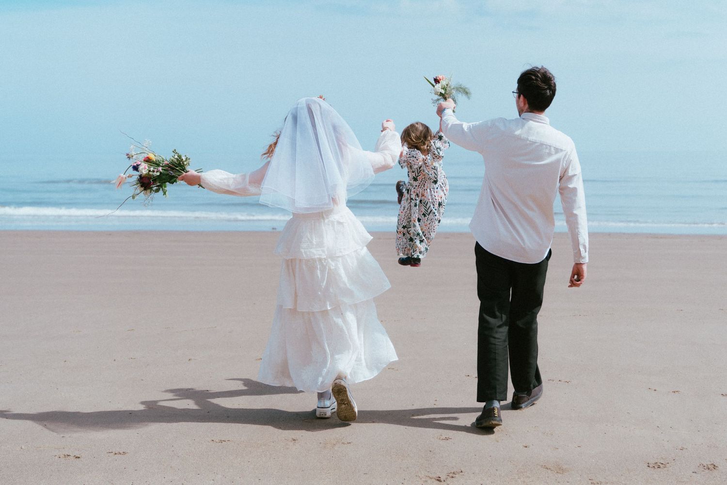 Bride in a white veil and gown and groom in a white shirt walk hand in hand with a small child along a sunny beach, holding flower bouquets.