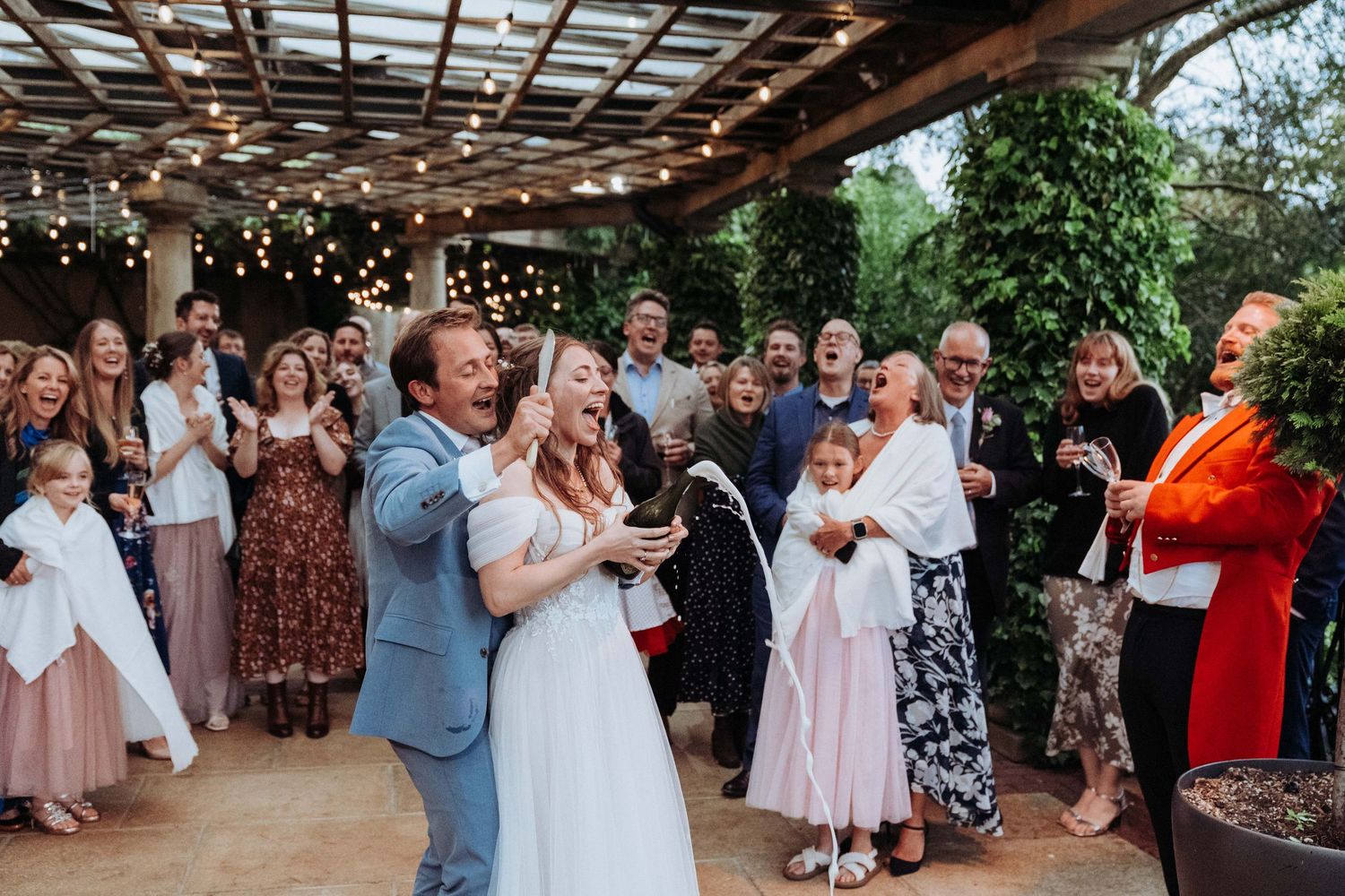 Bride and groom celebrate with guests as champagne bottle is opened at outdoor wedding reception.