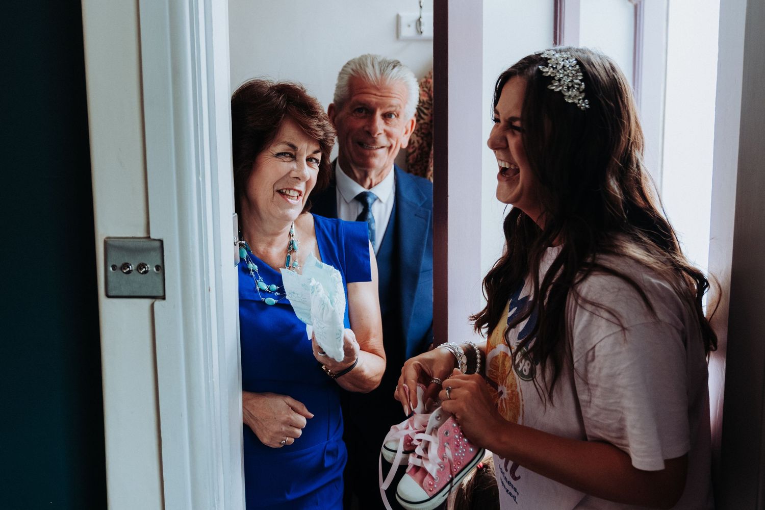 Bride in white top laughing with older couple at doorway during wedding morning preparations.