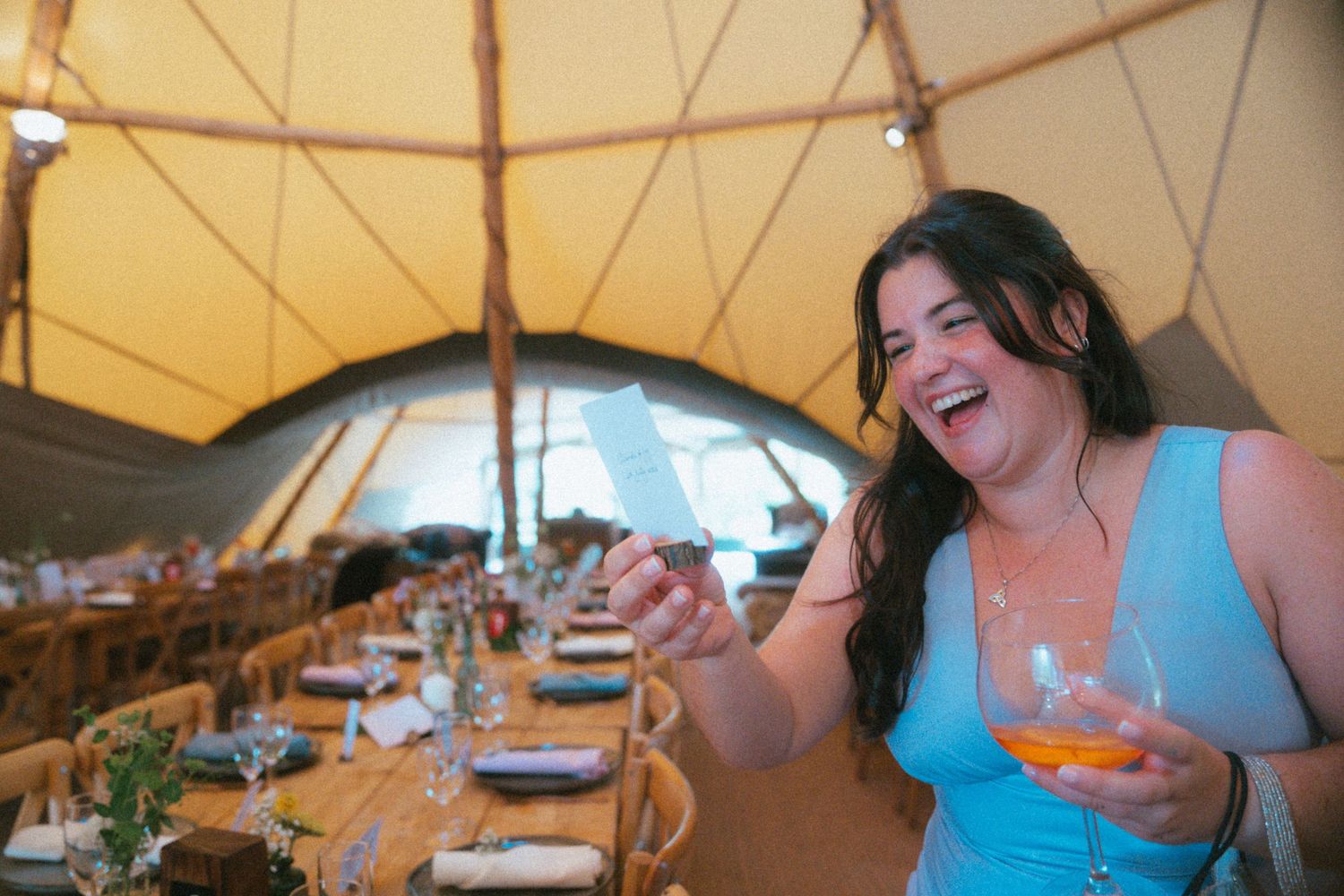 Happy woman laughing inside a tipi wedding reception tent, holding a drink at a decorated dining table.