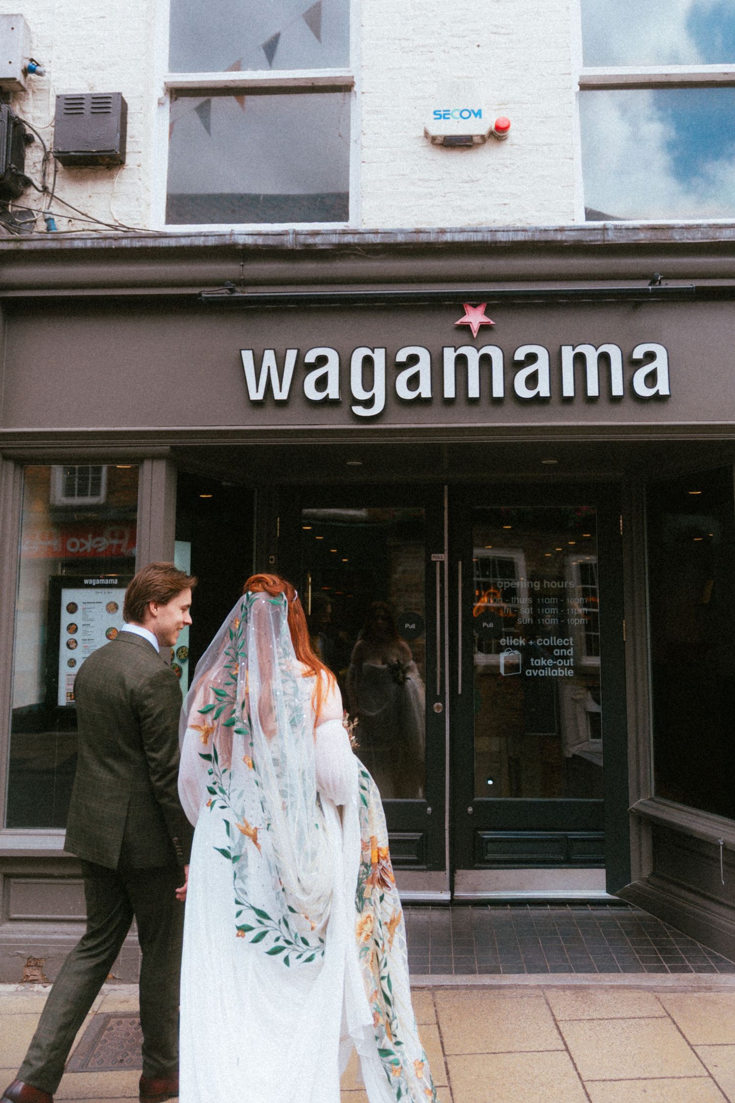 Bride in floral veil and groom walk toward Wagamama restaurant entrance on their wedding day.