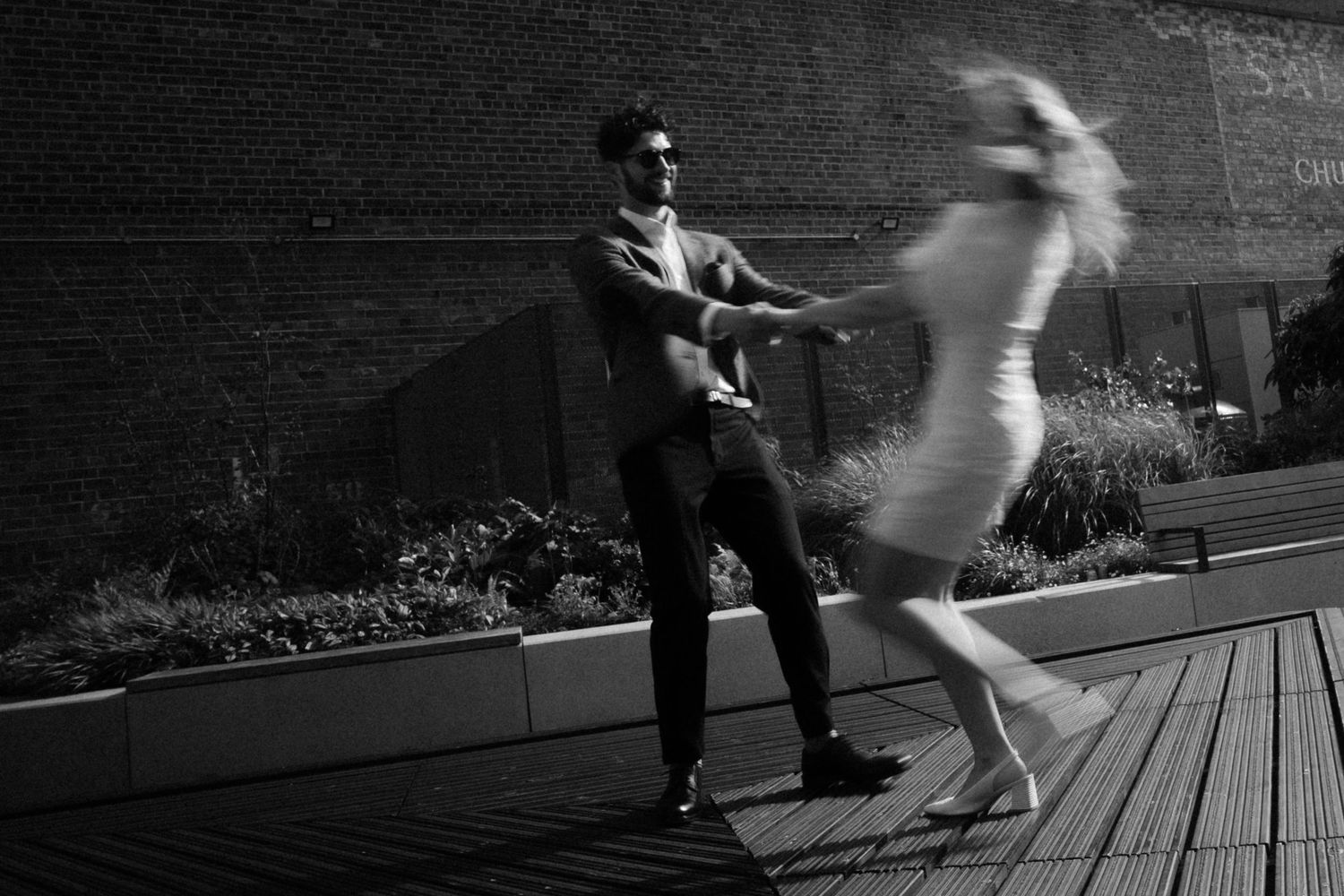 Black and white photo of a couple dancing outdoors on a wooden deck, with motion blur capturing their dynamic movement.