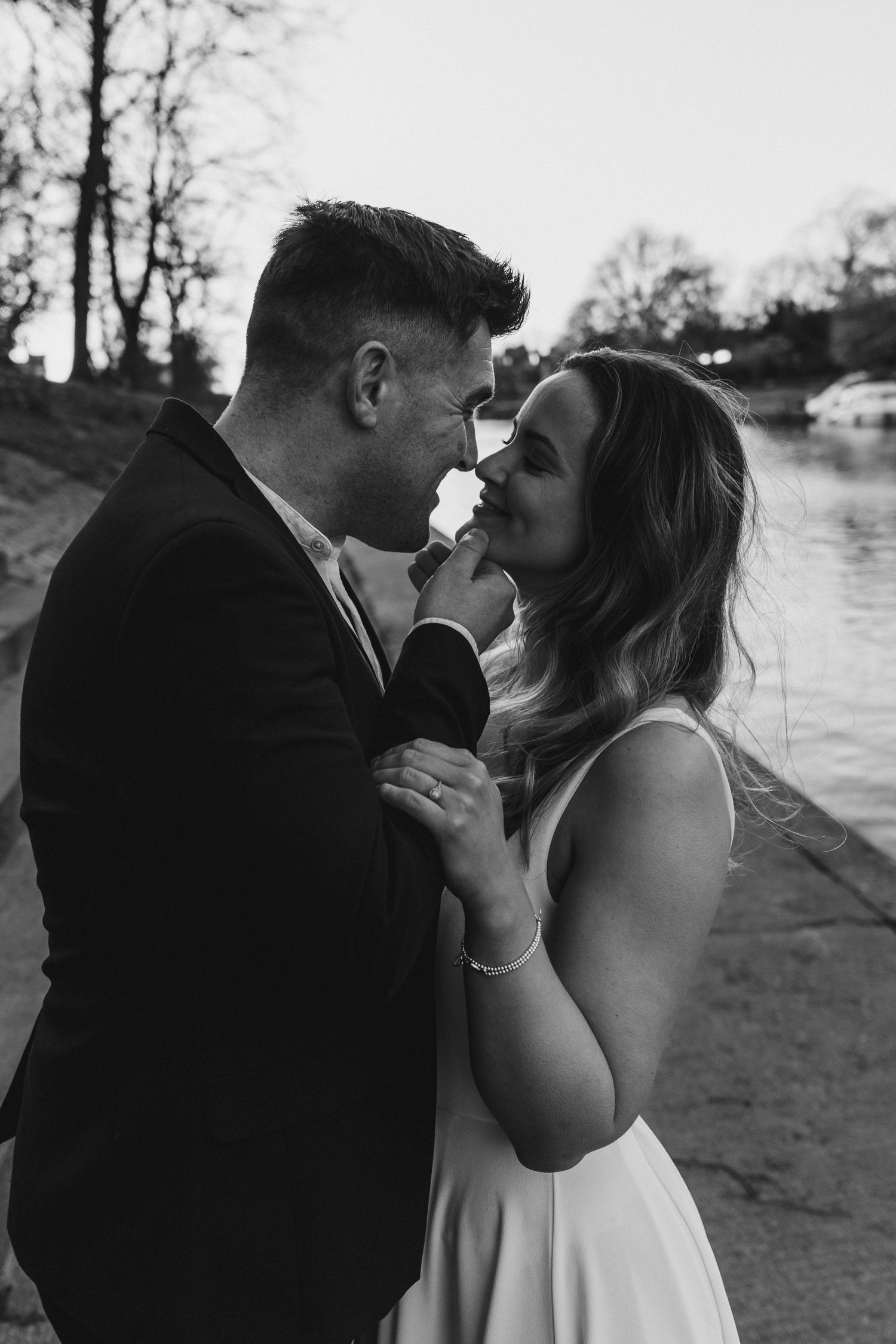 Bride and groom share an intimate moment near a river at dusk in black and white.