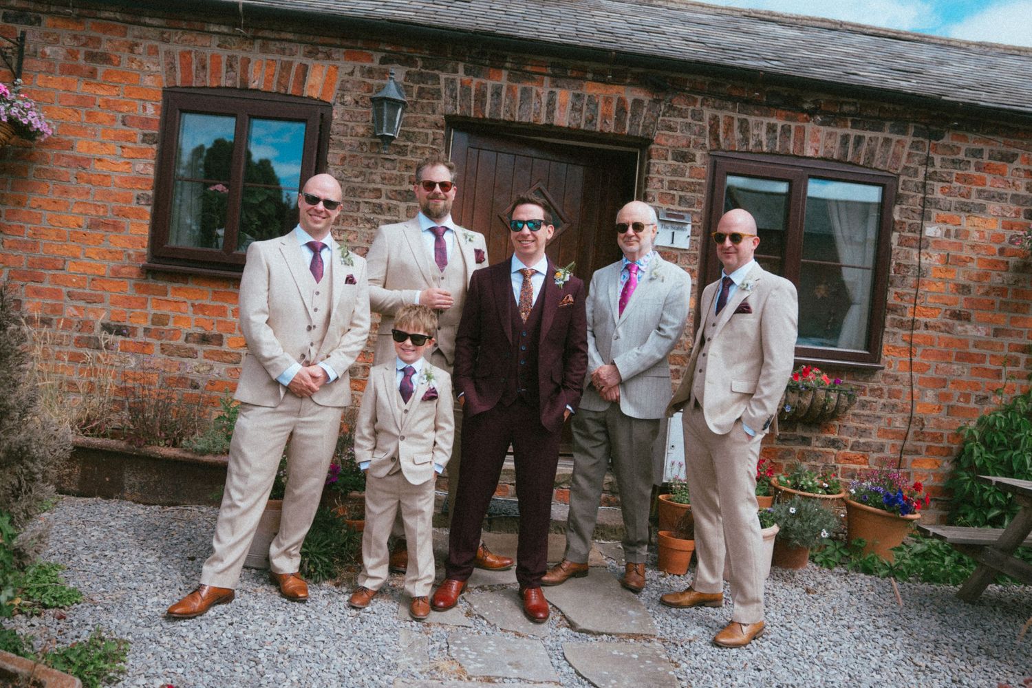 Groom in dark suit with groomsmen and young ring bearer in matching tan suits posing outside rustic brick wedding venue.
