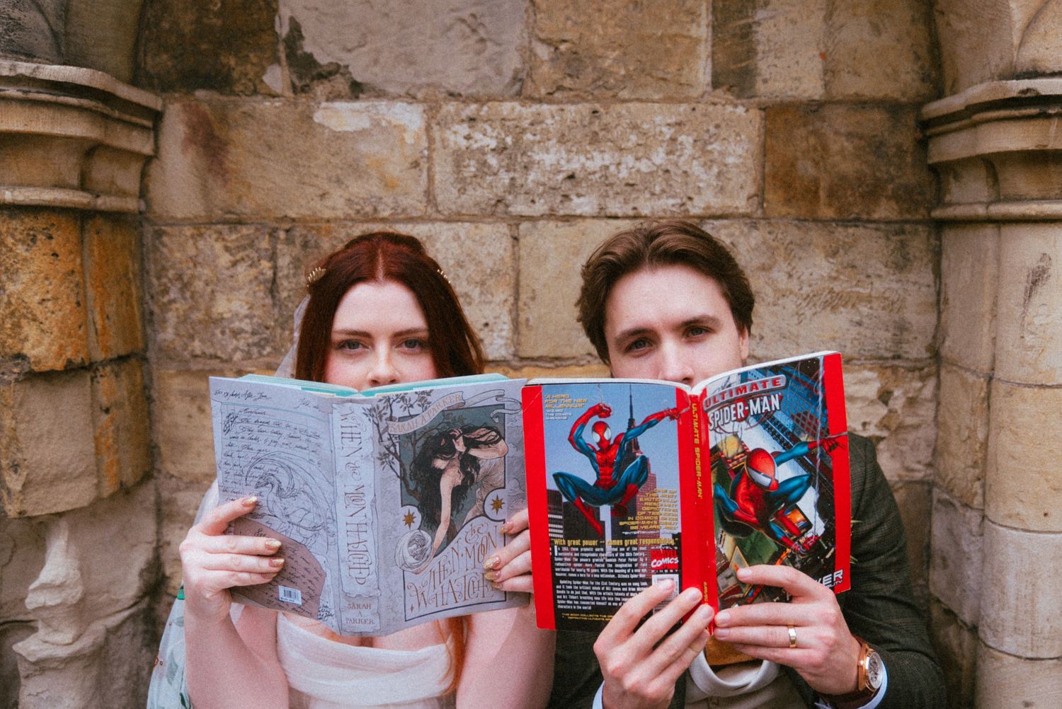 Bride and groom hiding faces behind Spider-Man comic books outside stone church wall.