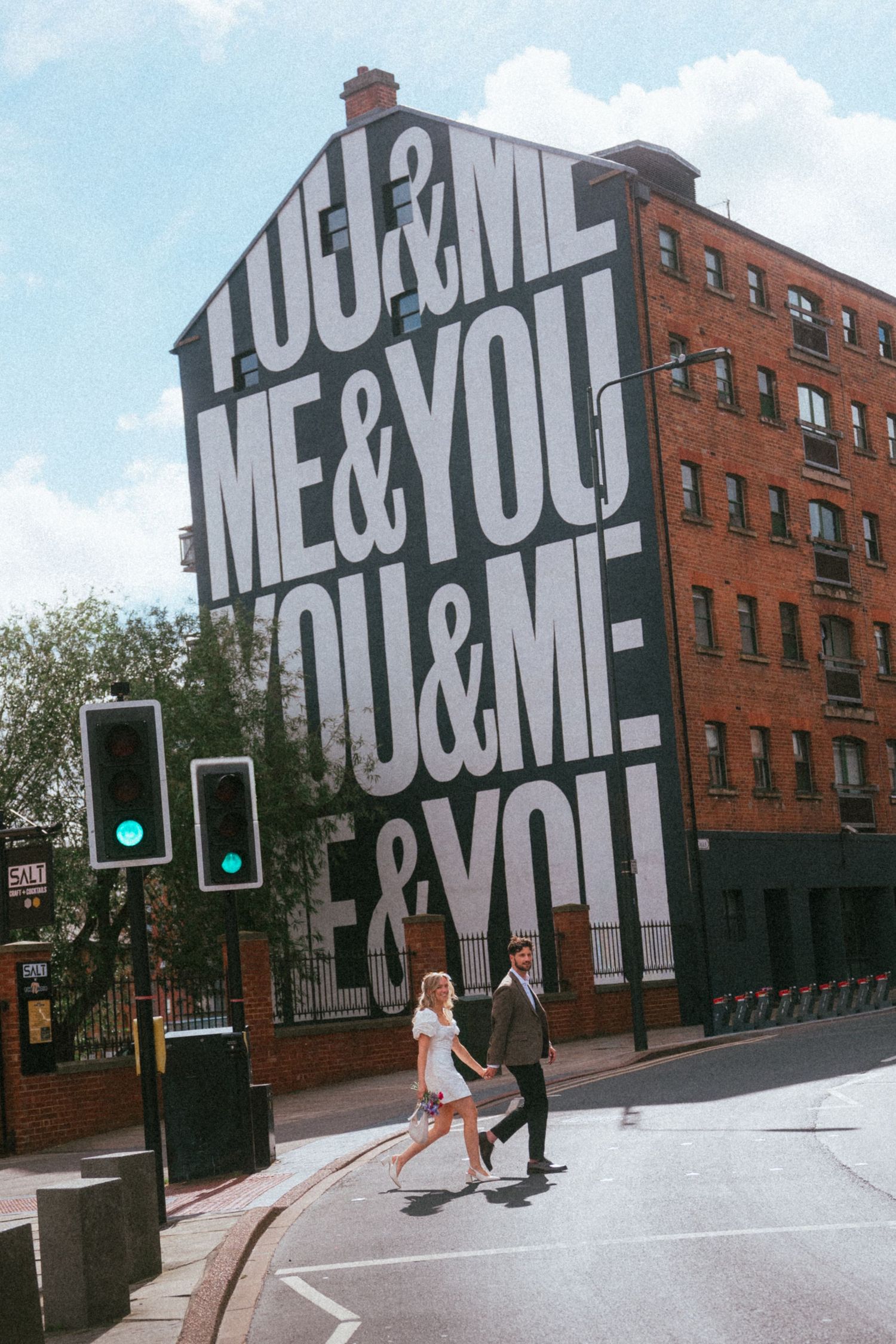 Couple crossing street in front of large YOU&ME mural on brick building in urban setting.