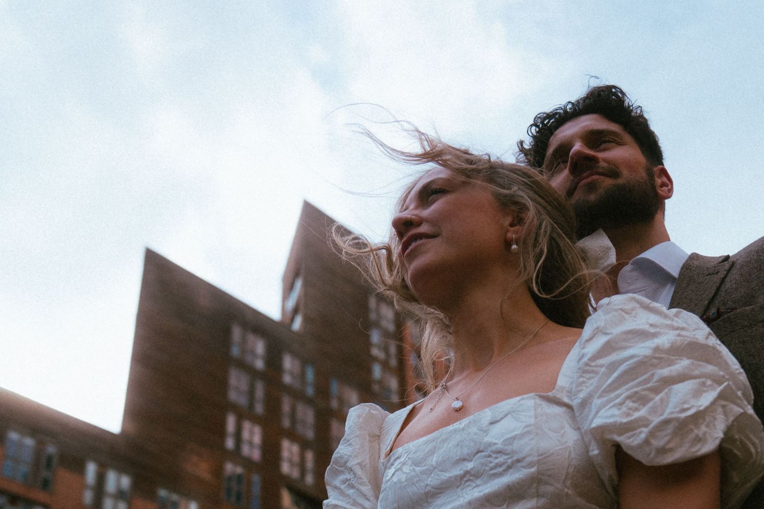 Bride and groom embracing outdoors with wind-blown hair against modern brick building and cloudy sky.