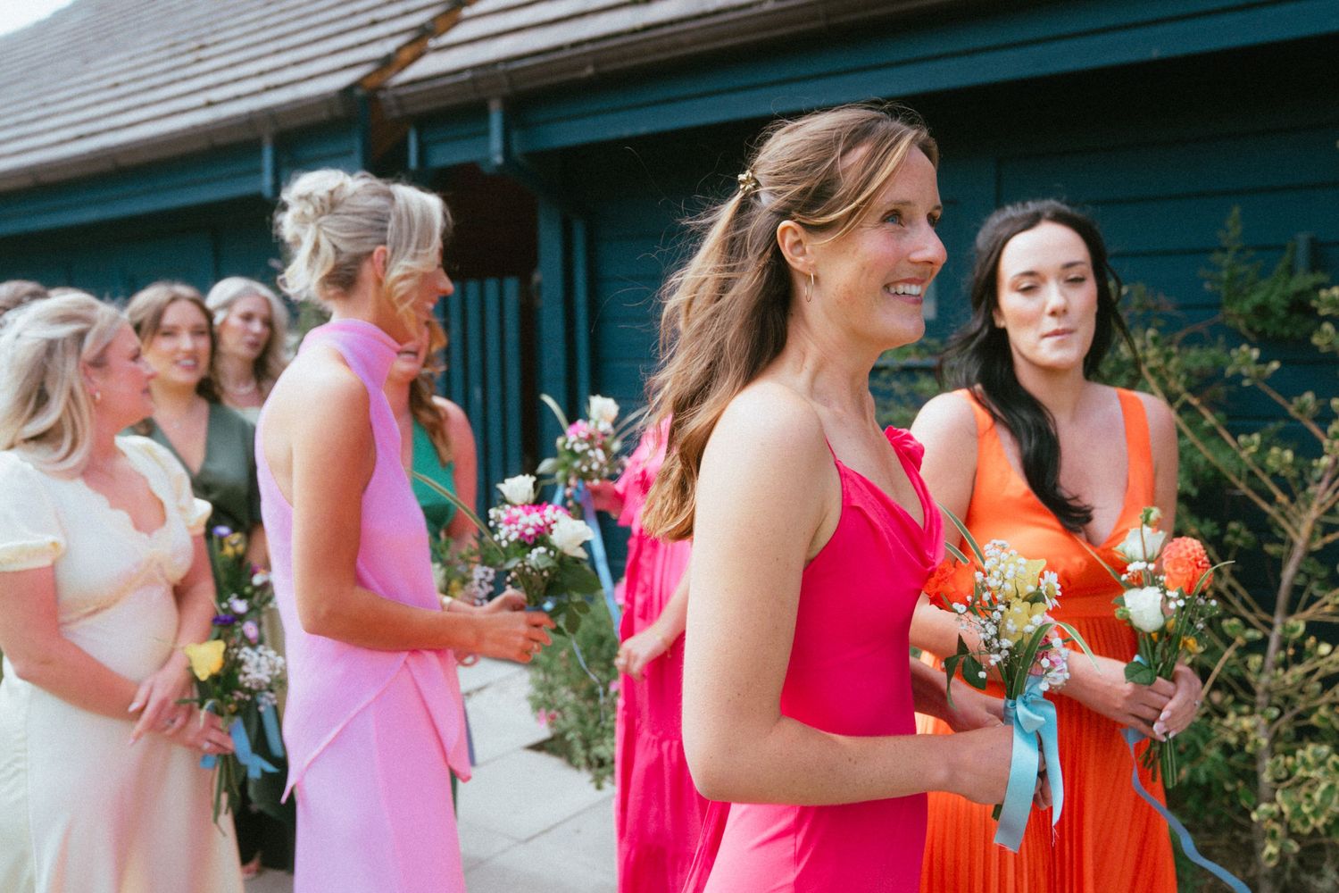 Bridesmaids in coral, pink and orange dresses hold colorful flower bouquets while smiling outside a rustic green wooden building.