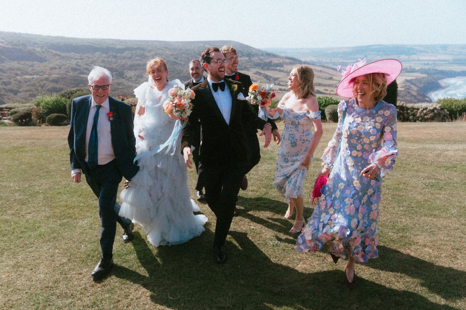 Bride in white gown and groom in tuxedo walk with smiling wedding guests across a sunny grassy hilltop overlooking the sea.