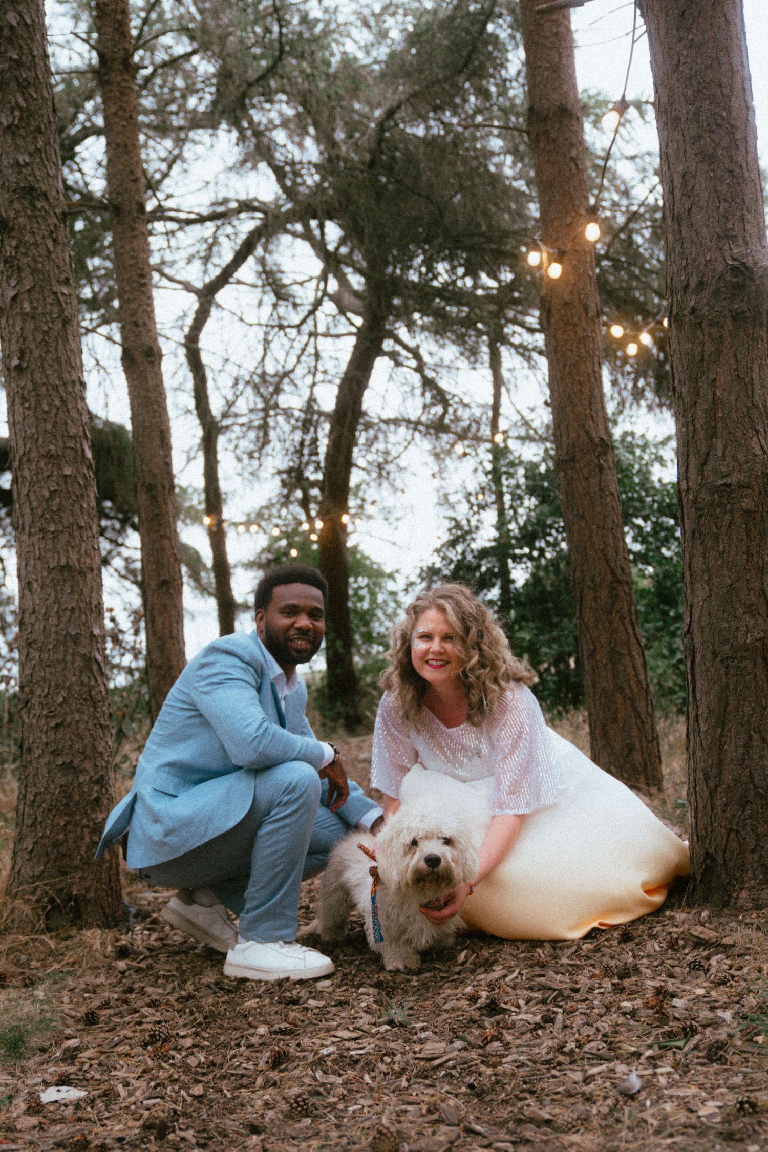A couple in wedding attire crouches with a white dog among trees with string lights in a forest setting.