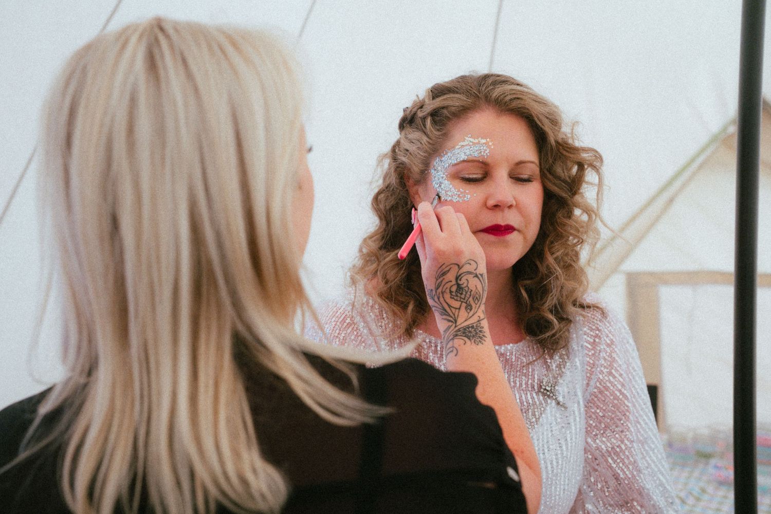 A blonde woman applying colorful face paint to a curly-haired woman near a white tent.