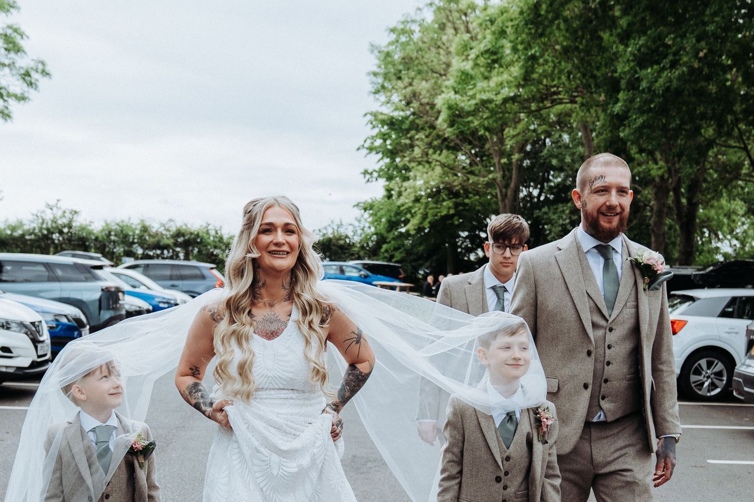 Bride and groom smiling with veil blowing in wind, accompanied by children in matching grey suits.