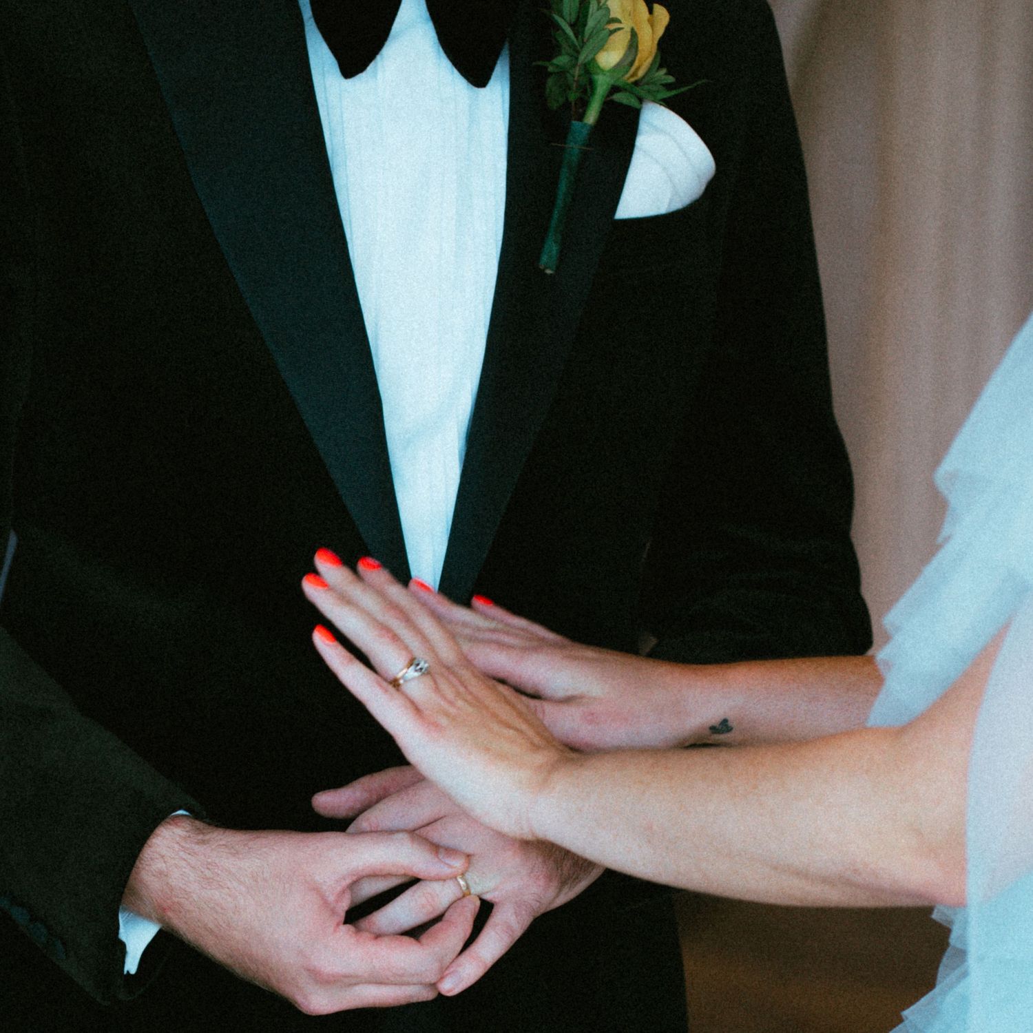Close-up of a groom in a black tuxedo with a yellow boutonniere as the bride's hand with red nails and a ring rests on his.