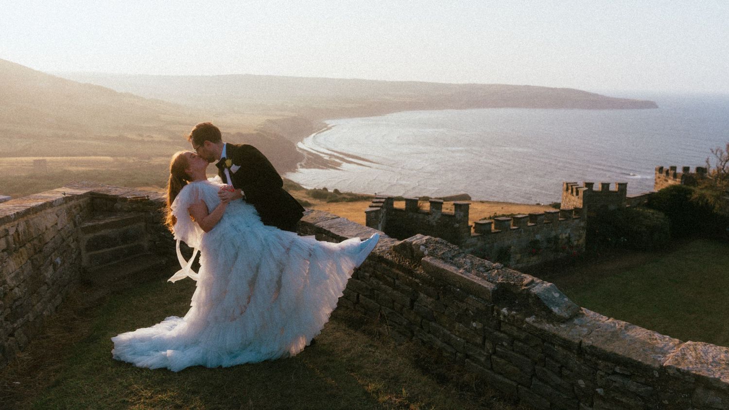Groom dips bride in a flowing white wedding gown on a stone castle wall overlooking a scenic coastline at golden hour.