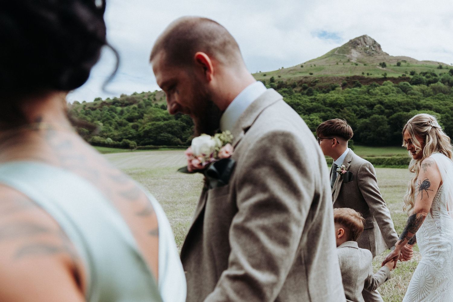 Groom in grey tweed suit with boutonniere walking with wedding party in scenic green countryside.