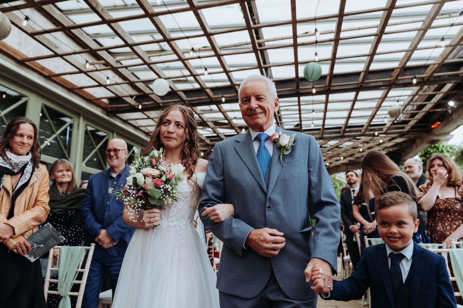 Bride walking down the aisle with father and young ring bearer at outdoor wedding.