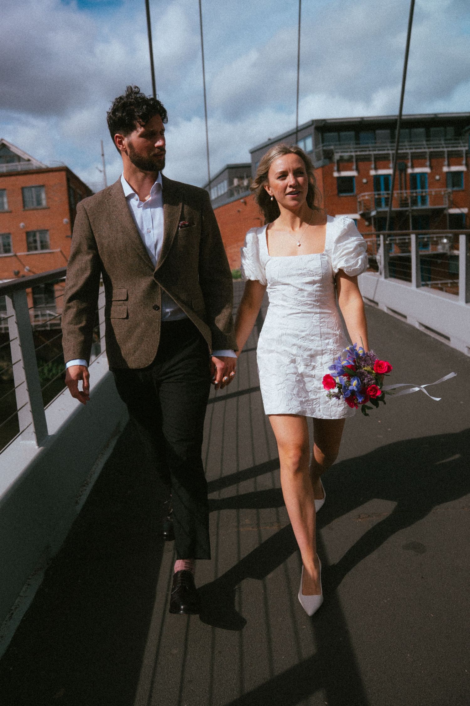 Bride in white mini dress and groom in brown suit walk hand-in-hand across a bridge on their wedding day.