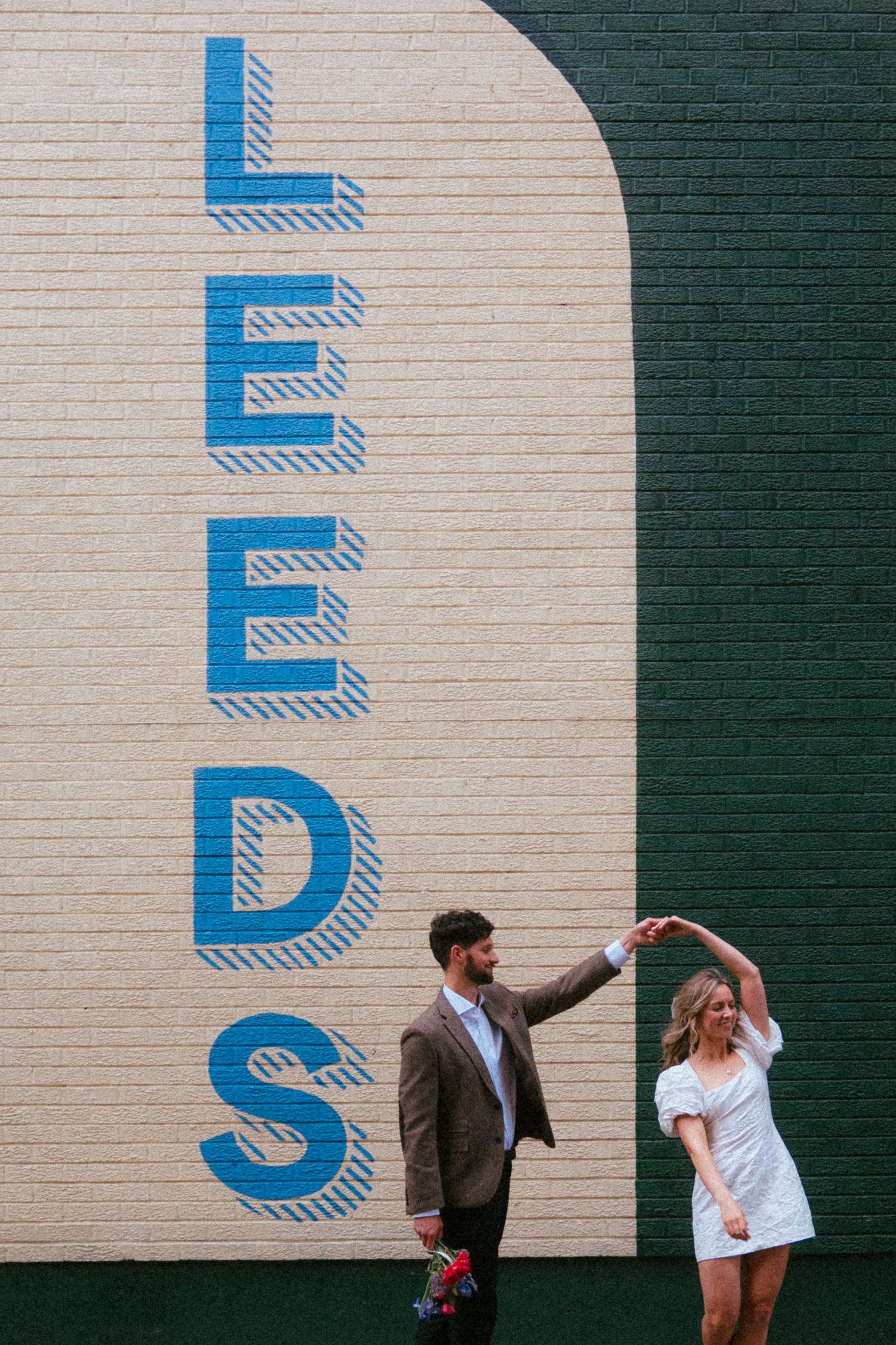 Couple dances playfully in front of a large LEEDS mural painted on a cream and green wall.