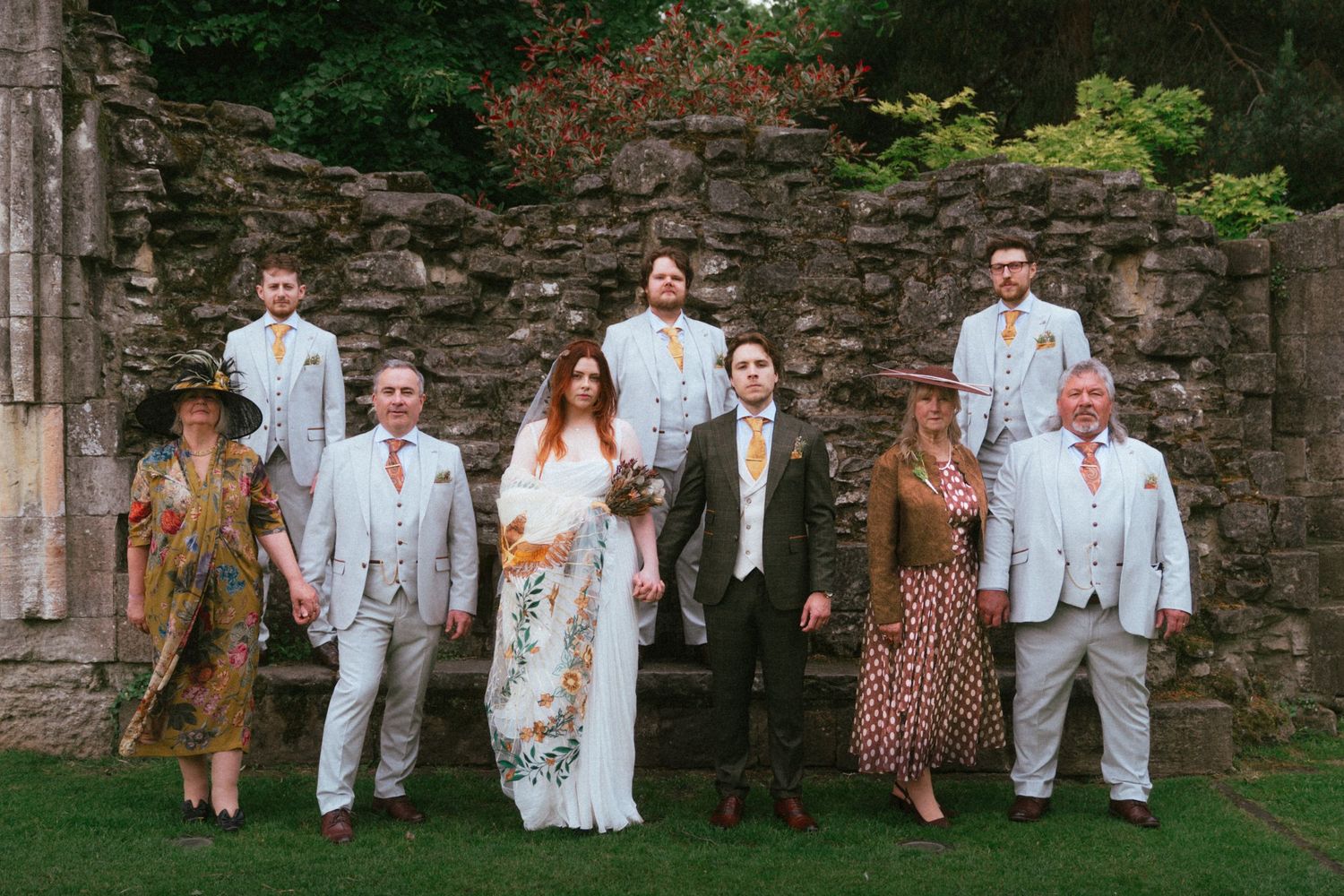 Wedding party group portrait in front of rustic stone ruins, bride in boho floral dress with groom in tweed suit.
