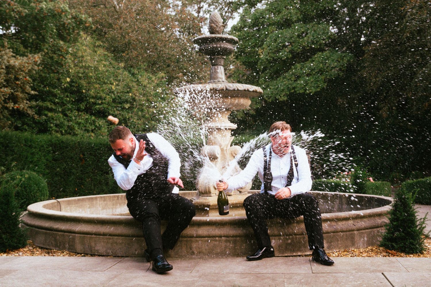 Two men in matching vests and bow ties crouch beside a stone garden fountain, spraying champagne in a joyful wedding celebration.