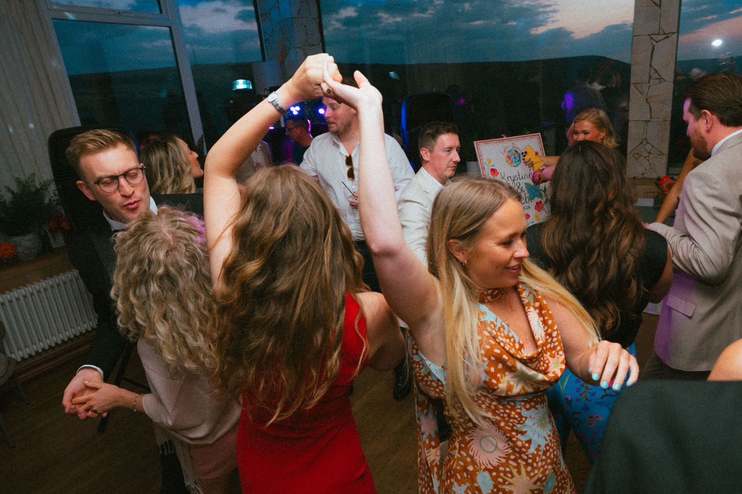 Wedding guests dance and raise their arms together on a lively dance floor in a venue with large windows at dusk.