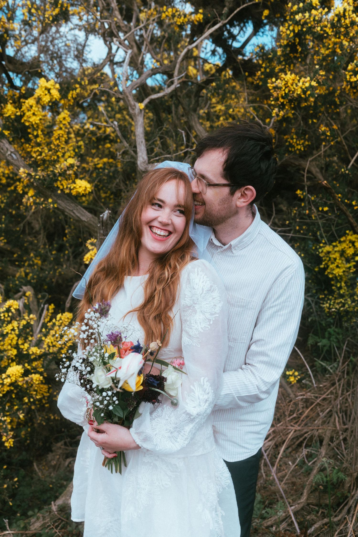 A smiling bride in a white lace dress and veil holds a wildflower bouquet as her partner embraces her beside blooming yellow gorse.