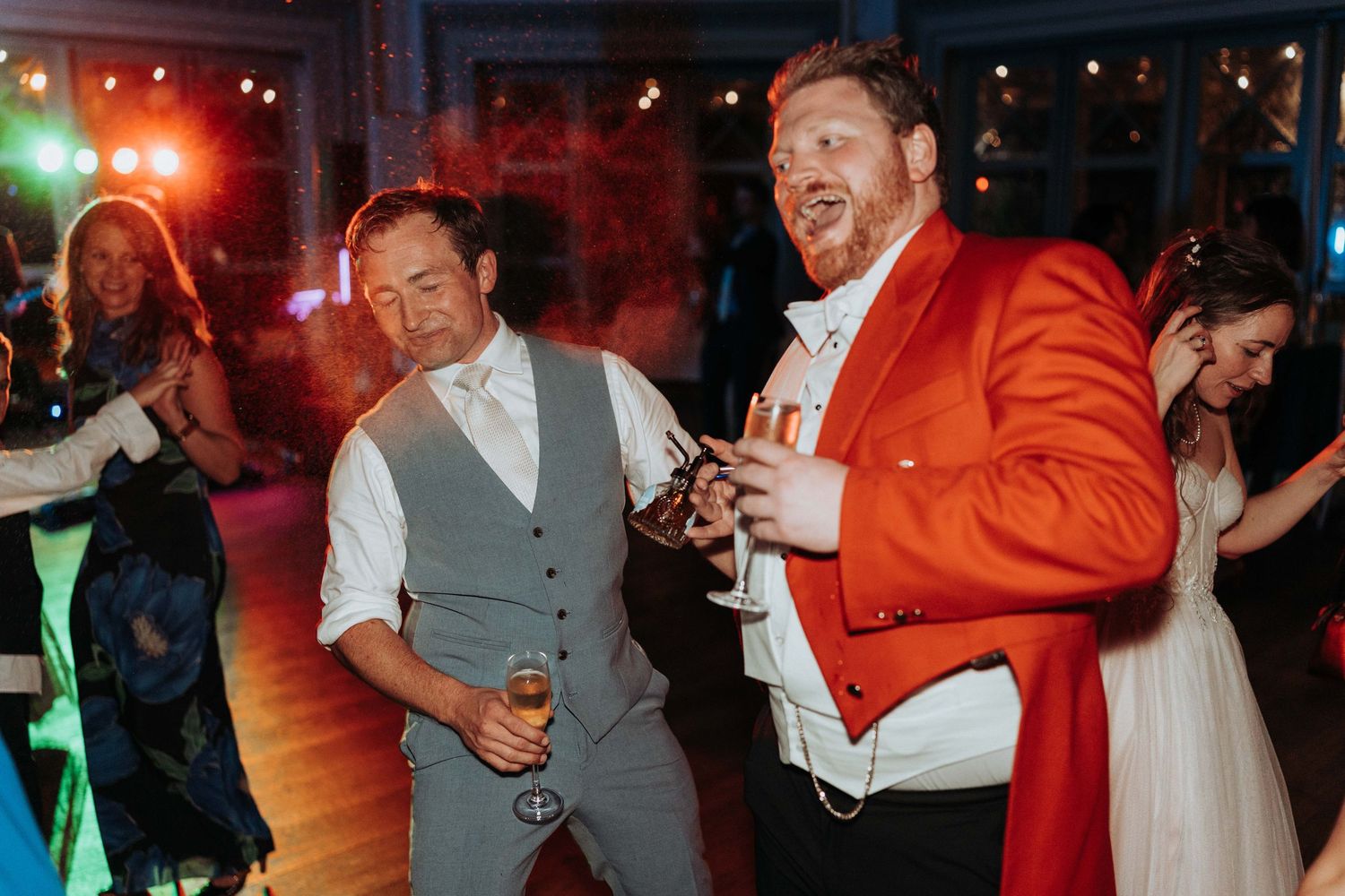 Two men celebrating and laughing on the wedding dance floor, one wearing a bold red jacket.