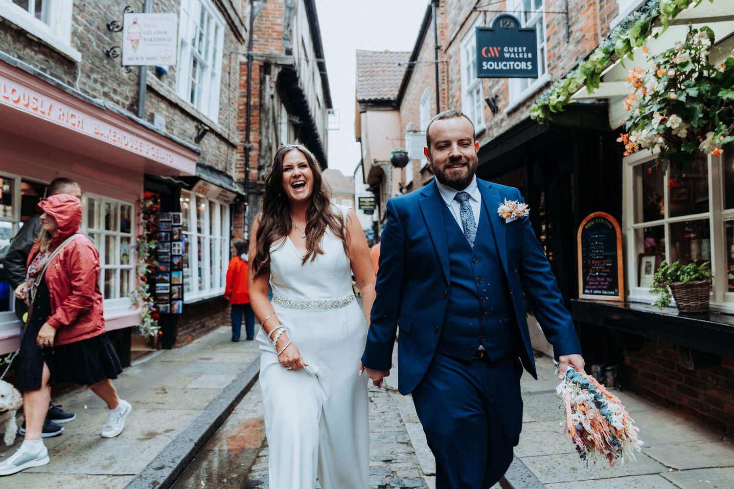 Laughing bride in white gown and groom in navy suit walk through charming cobblestone street in York.