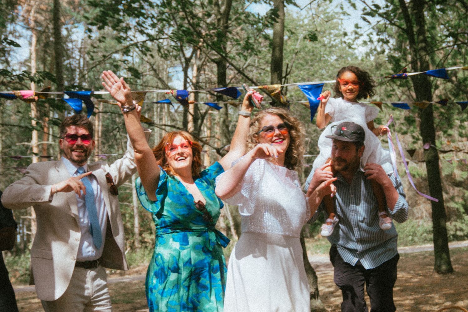 Joyful wedding party celebrating outdoors in a forest with colorful bunting, bride in white dress surrounded by guests.