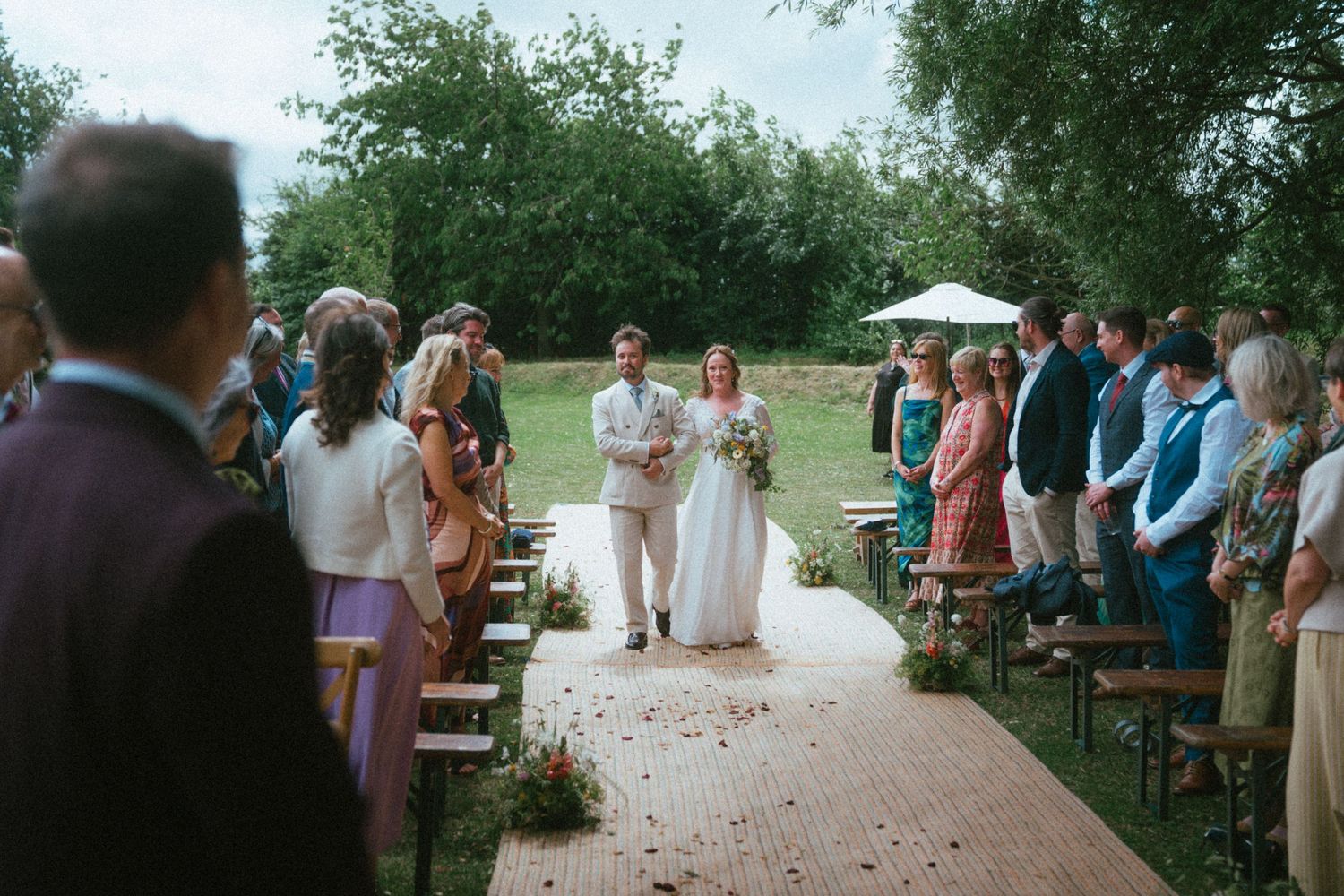 Bride and groom walk down outdoor wedding aisle surrounded by standing guests amid lush greenery.