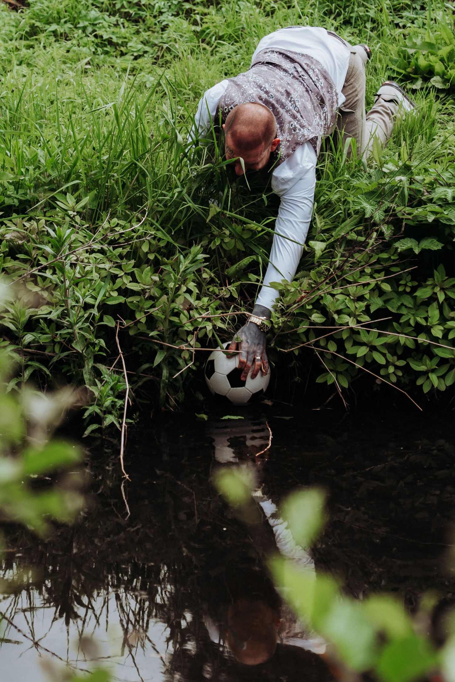 Tattooed groom in wedding attire reaches into a stream to retrieve a soccer ball from the water.