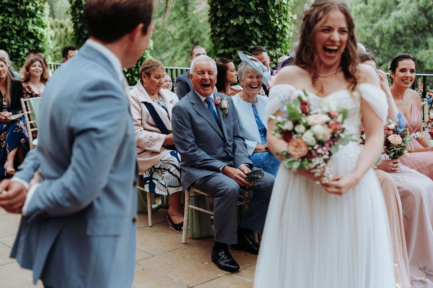 Laughing bride holding bouquet approaches groom as wedding guests smile and cheer during outdoor ceremony.