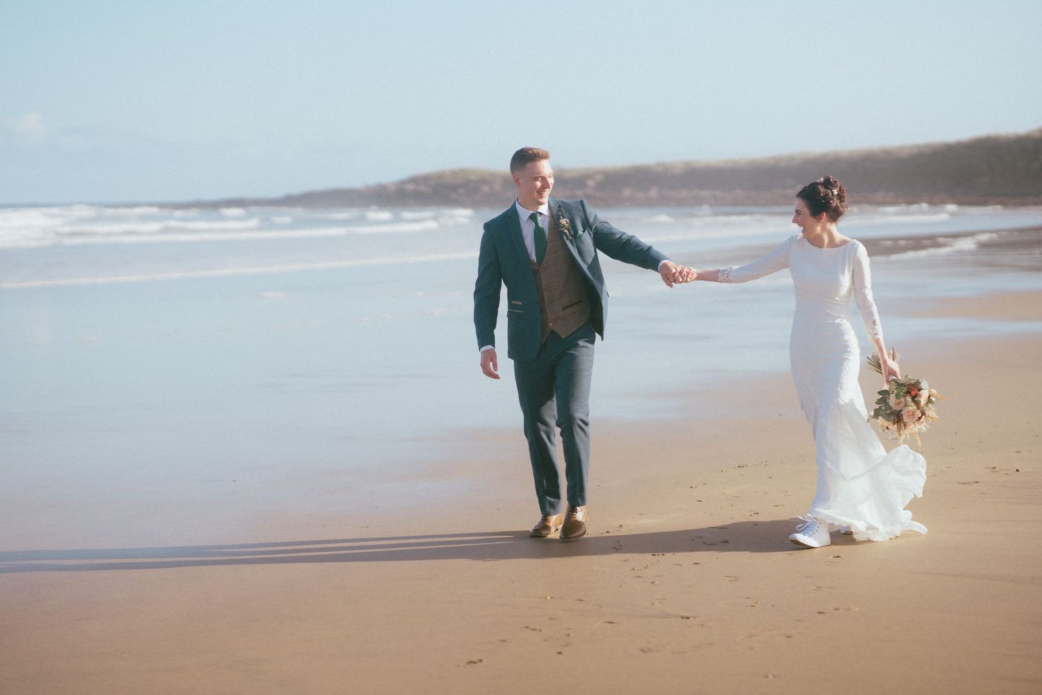 Bride and groom walking hand in hand along a sunny sandy beach with ocean waves in the background.